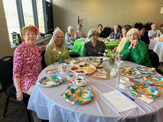 Group of elderly women celebrating at a table with colorful plates, teacups, flowers, and Easter-themed decorations in a room with large windows.