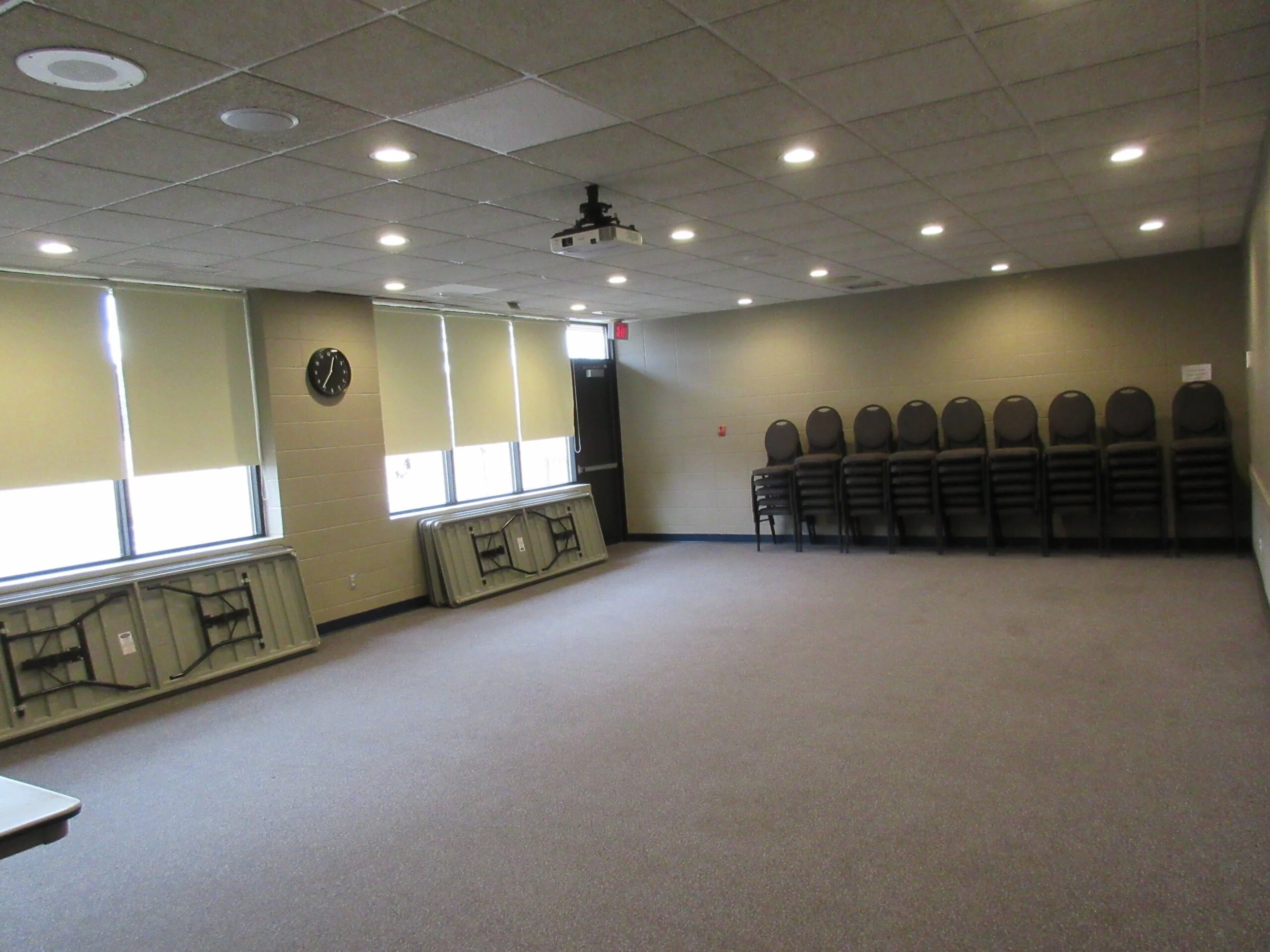 An empty room with a ceiling mounted projector, folding tables stacked on the floor, chairs stacked against the wall, a clock on the wall, windows with blinds, and carpeted floor.
