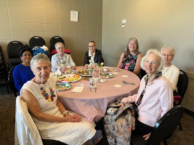 Six women sitting around a round table decorated with a pink tablecloth, plates of food, and a centerpiece with a bunny figure, in a room with gray walls and black chairs against the wall.