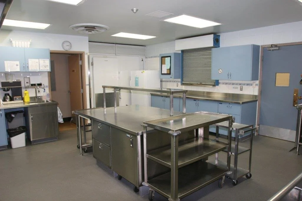 Commercial kitchen with stainless steel tables and cabinets, a white refrigerator, a no-smoking sign, and various kitchen outlets and appliances.
