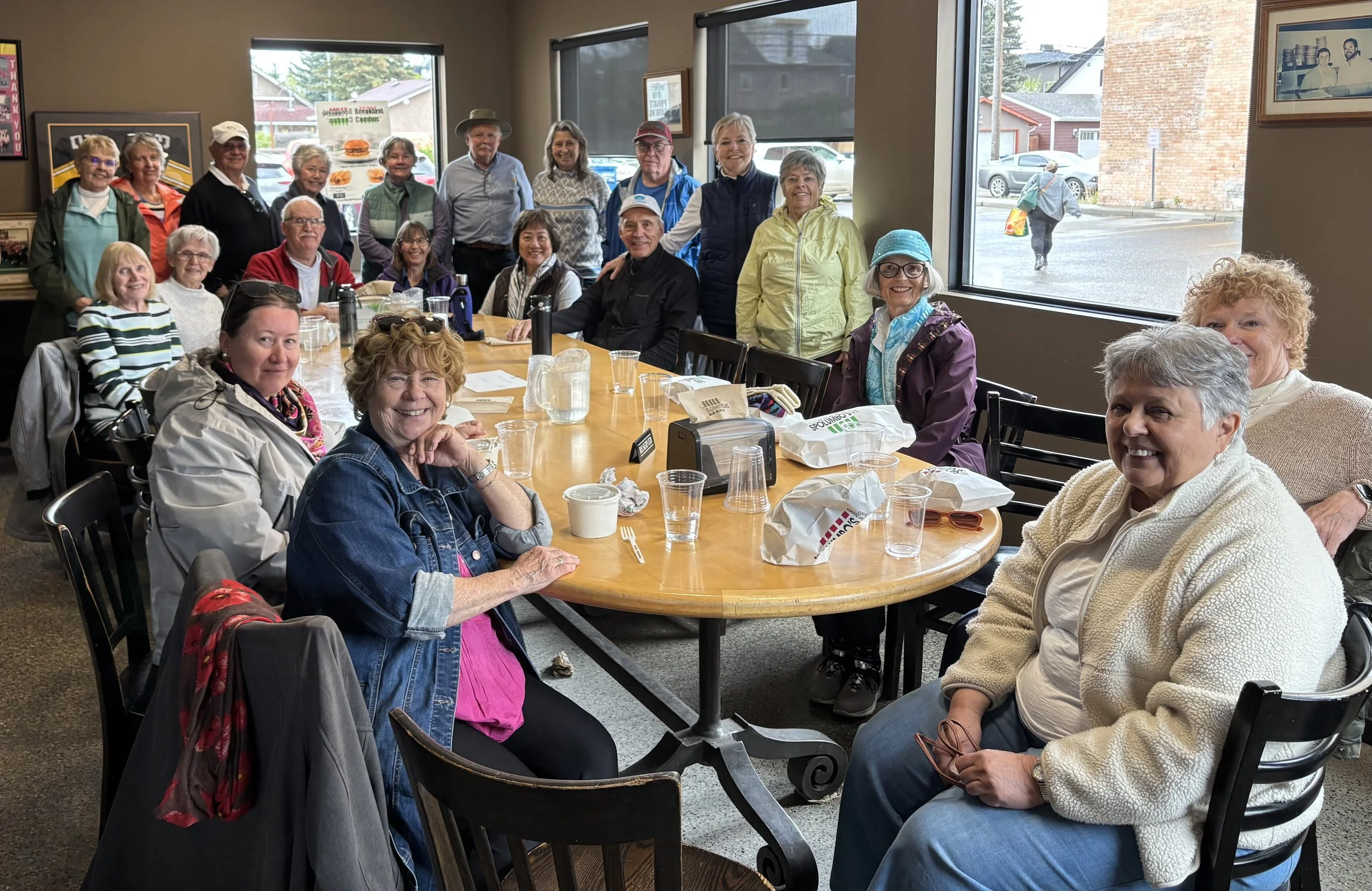 A group of elderly people gathered around a large wooden table inside a restaurant. Some are sitting, others standing behind the seated ones. The table is set with cups, glasses, and paper bags. Outside the window, a woman with a colorful bag walks across the rainy street. The inside has framed photos and posters on the walls.