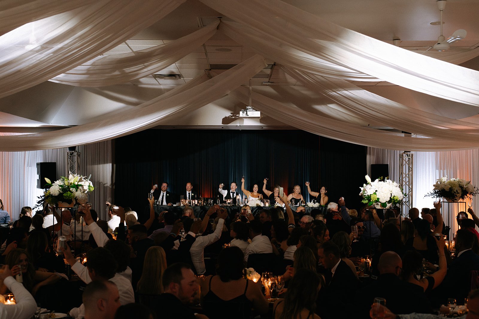 A wedding reception ceremony inside a decorated hall with draped fabric ceiling and floral centerpieces. The bride and groom, along with several guests, are raising glasses in celebration at a long head table on a stage with a black curtain backdrop. The room is filled with seated guests.