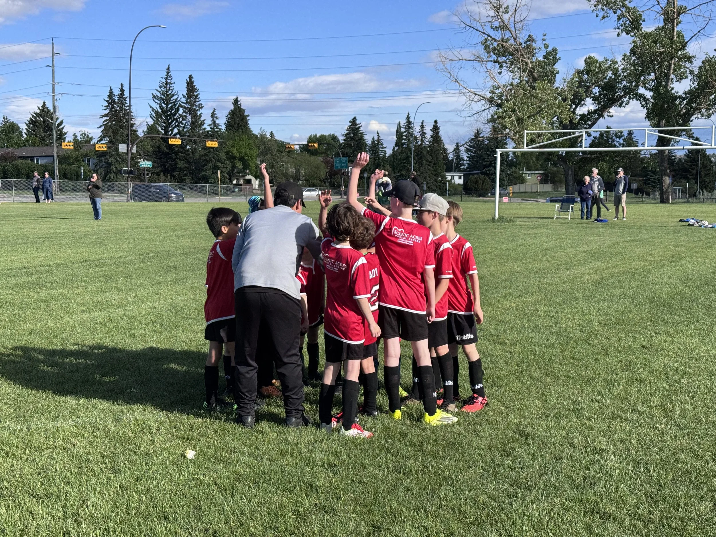 A youth soccer team wearing red jerseys standing in a line on a grassy field, with some raising their hands. The coach or referee is talking to them. In the background, there are spectators, trees, and a fence.
