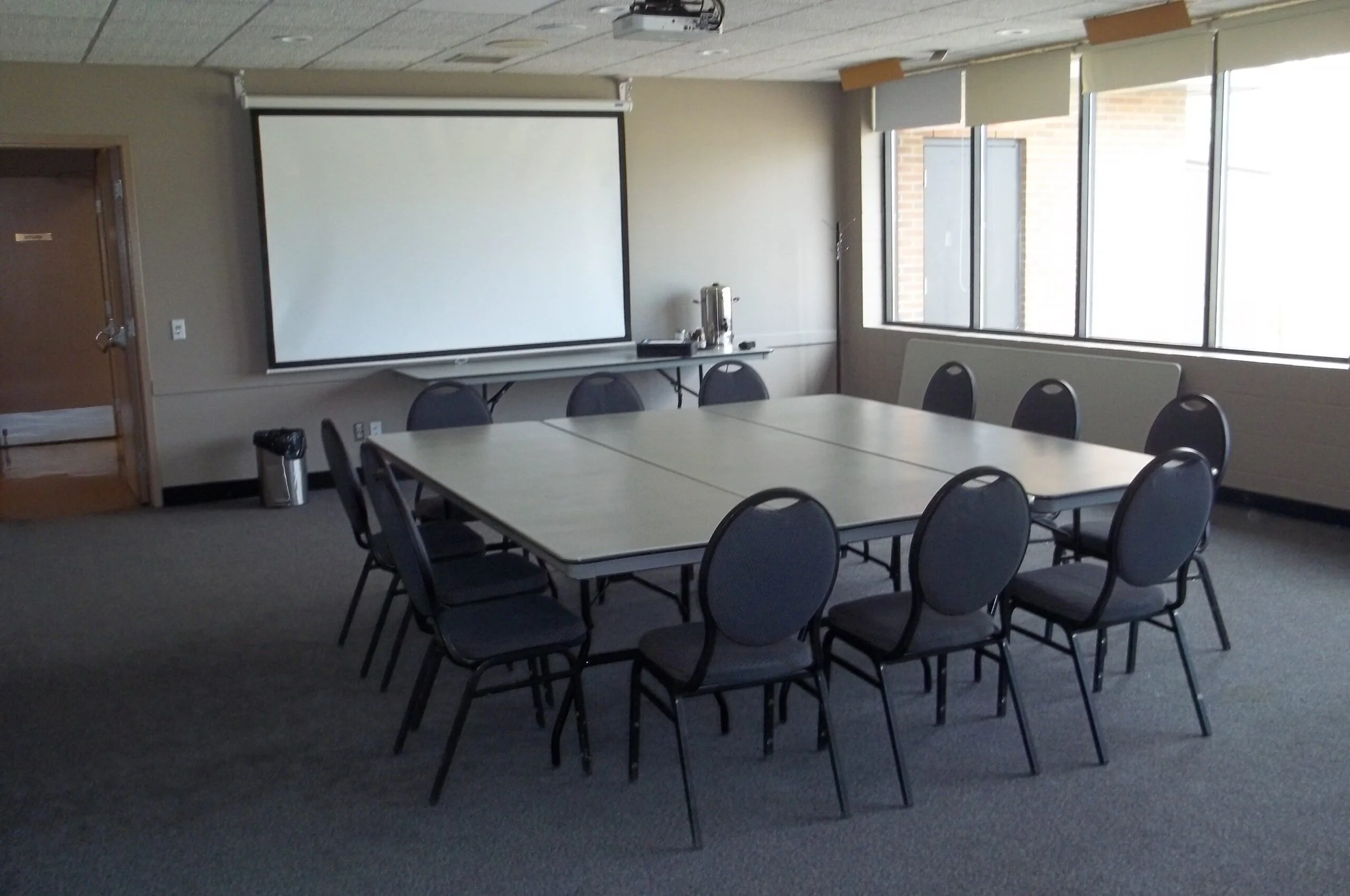A conference room with rectangular tables arranged in a U-shape. There are black chairs around the tables. A large white projection screen is mounted on the wall, with windows on the right side of the room allowing natural light in.