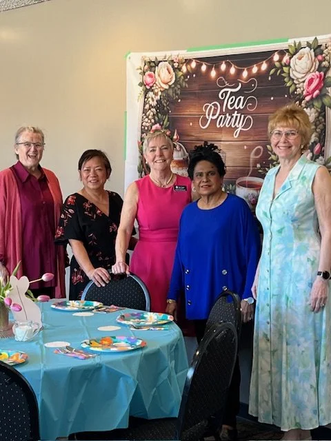 Five women standing together at a tea party event, decorated with floral and fairy lights backdrop, in a room with tables set with plates, utensils, and a centerpiece with balloons.