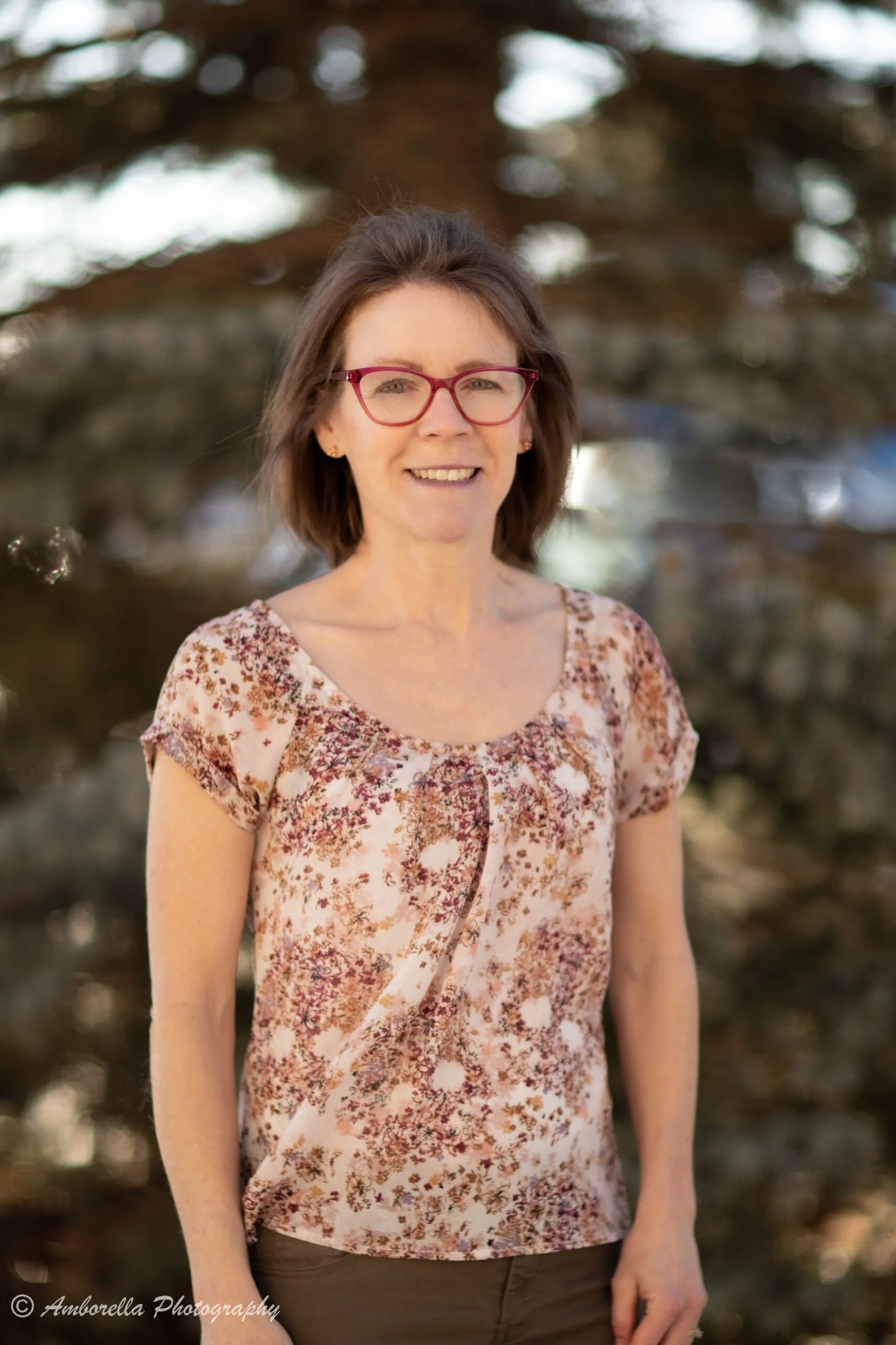 A woman with short brown hair and glasses smiling outdoors near a large tree, wearing a floral patterned blouse and dark pants.