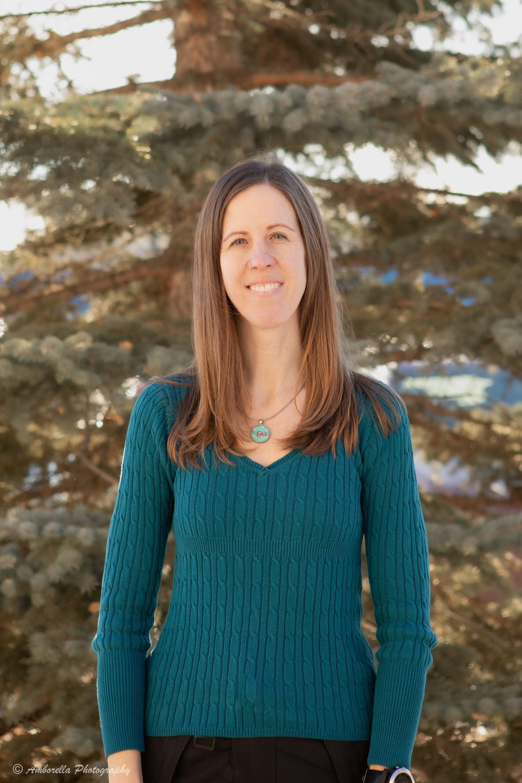 A woman with long brown hair wearing a teal cable-knit sweater and a necklace with a poppy pendant, standing outdoors with a background of large evergreen tree branches.