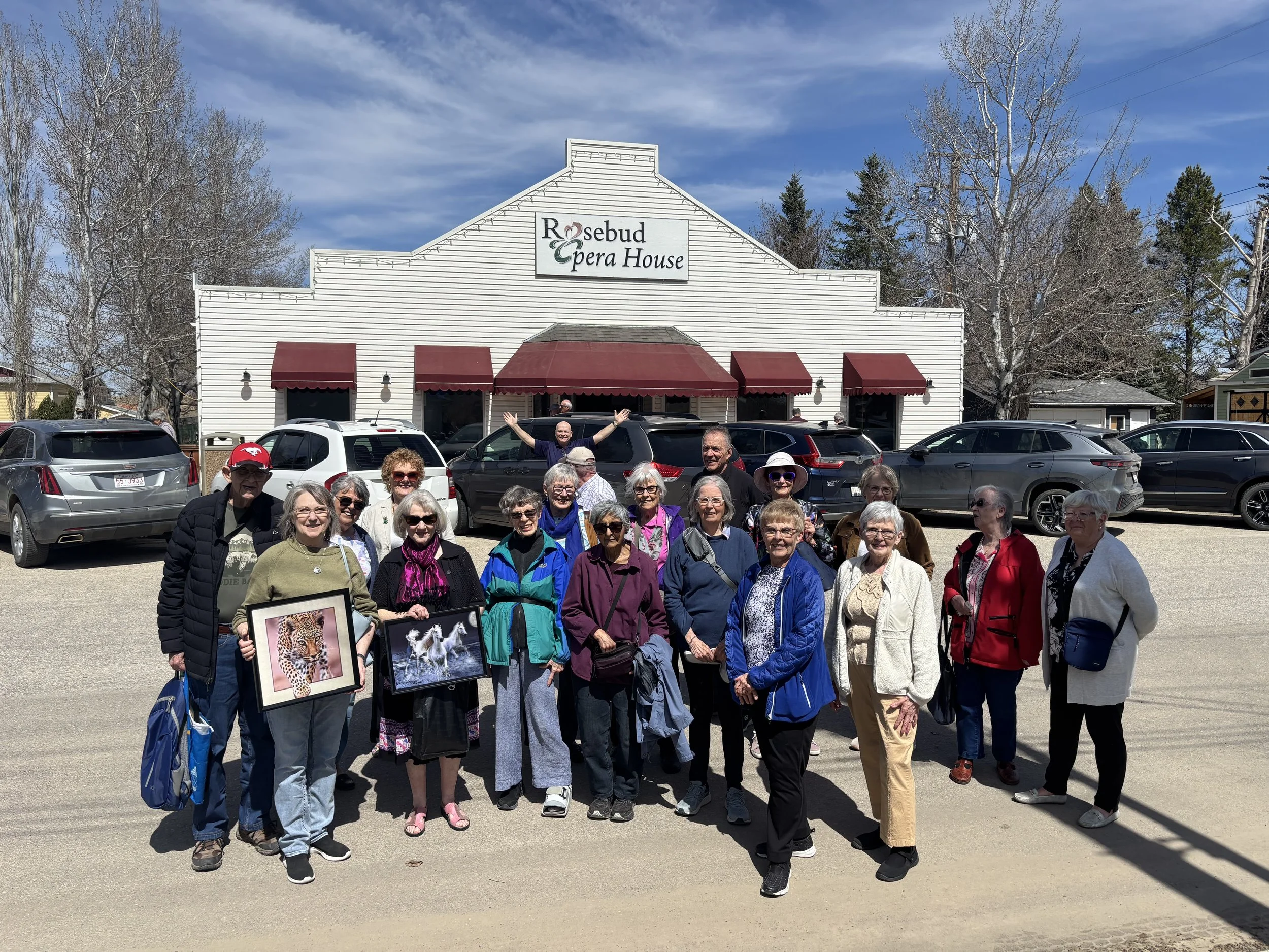 Group of older adults standing in front of the Rosebud Pera House, some holding framed pictures of animals, on a parking lot with parked cars and leafless trees against a partly cloudy sky.