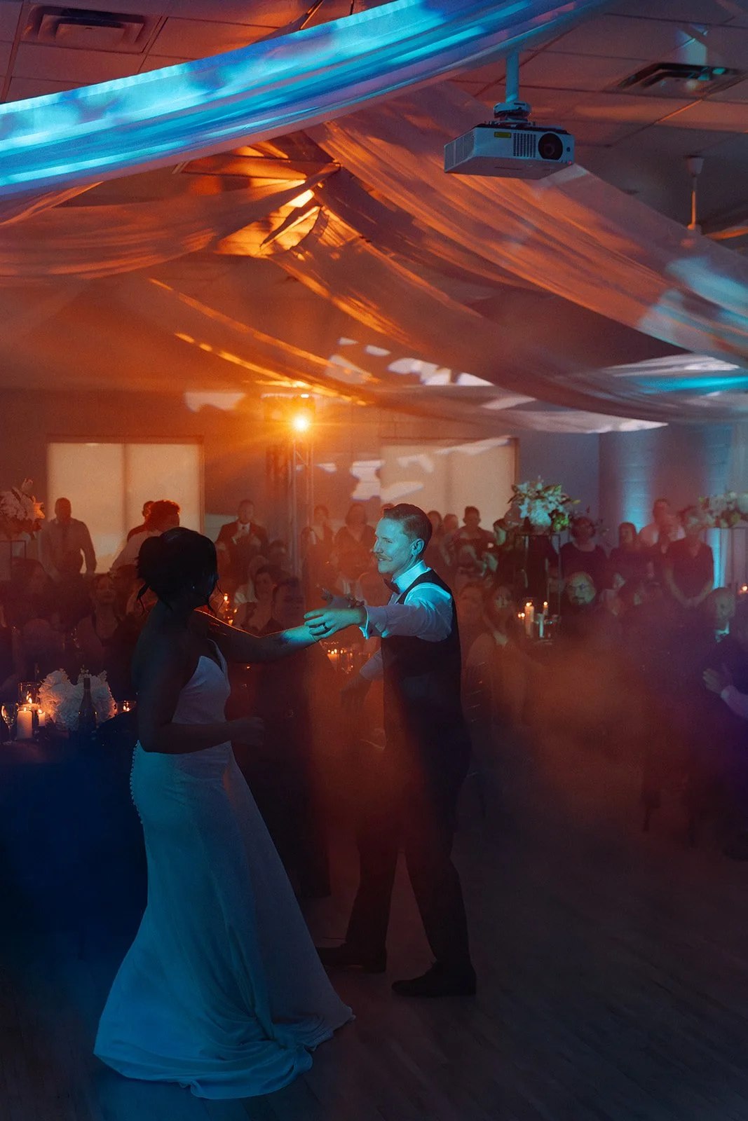 A couple dances at a wedding reception, surrounded by guests, with colorful drapes and lighting overhead.