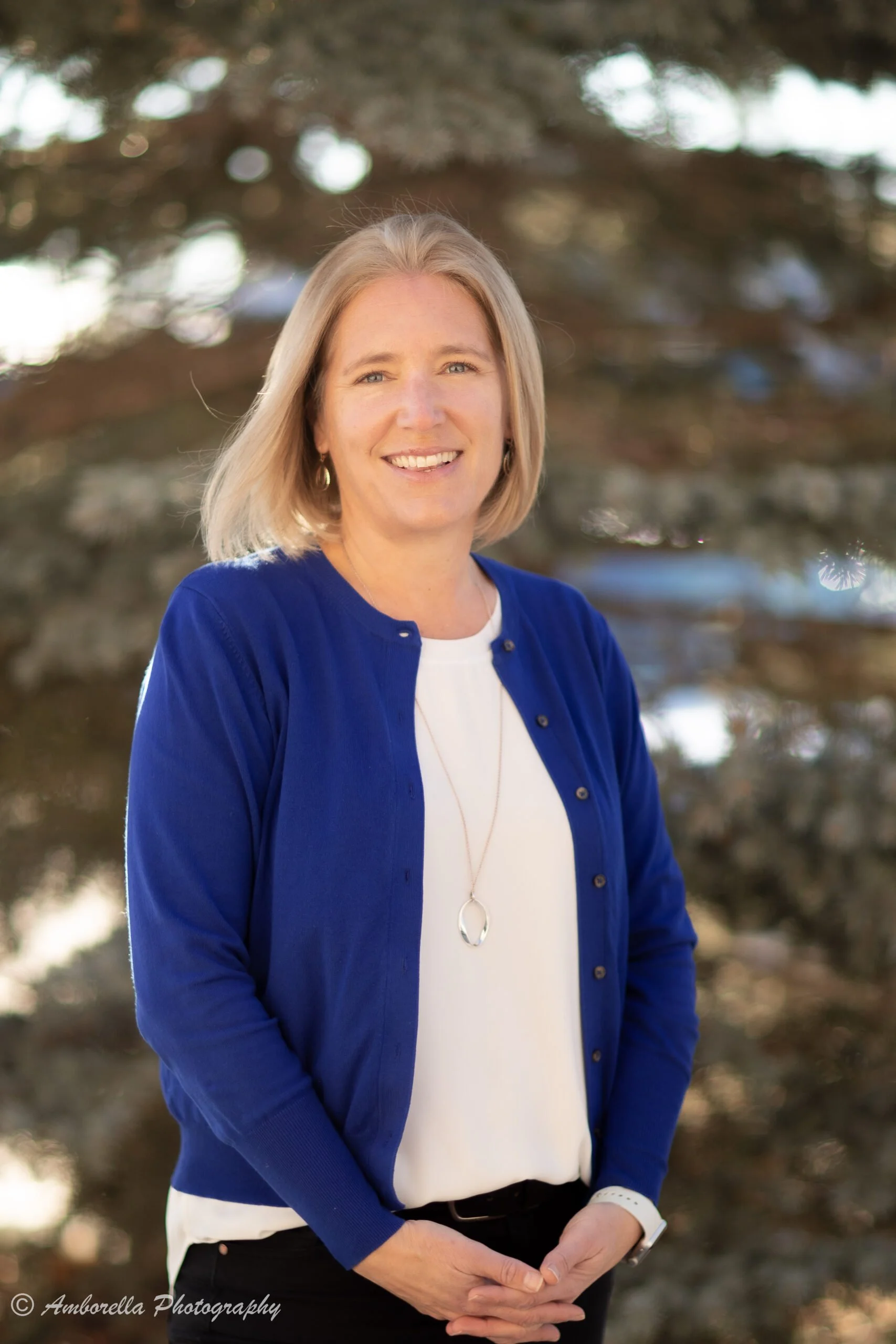 A woman with blonde hair wearing a blue cardigan over a white shirt, standing outdoors in front of trees, smiling at the camera.