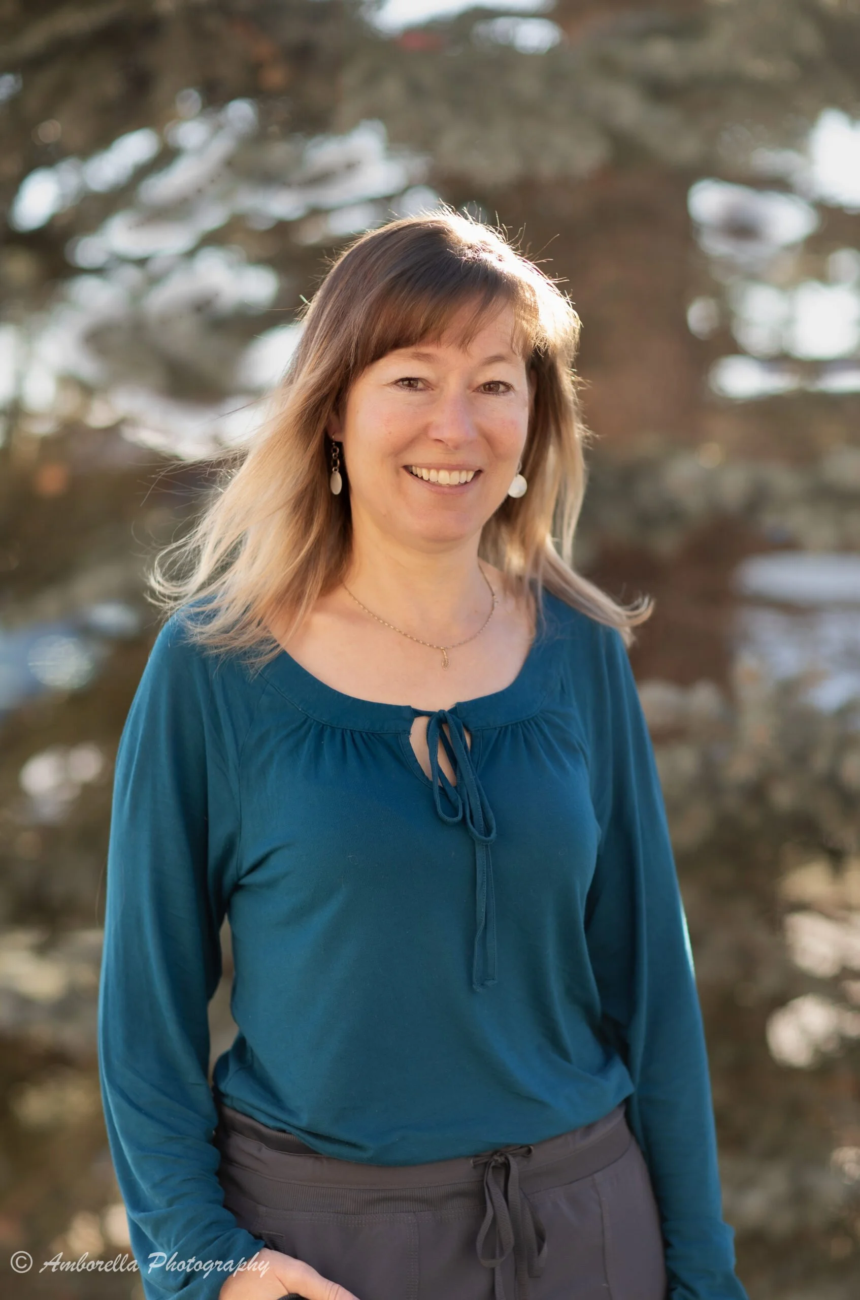 A woman with shoulder-length light brown hair, wearing a blue blouse and gray pants, standing outdoors in front of blurred trees, smiling at the camera.