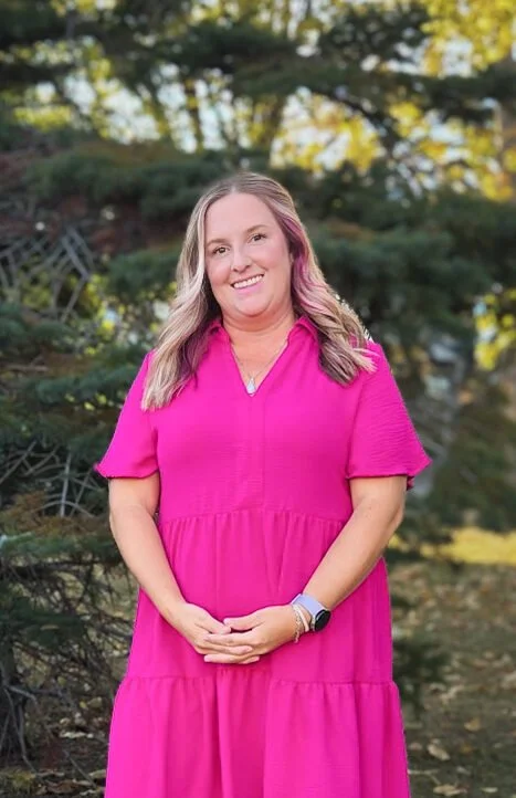A woman with long, wavy blonde hair and pink streaks, wearing a bright pink dress, standing outdoors in front of green trees and sunlight.