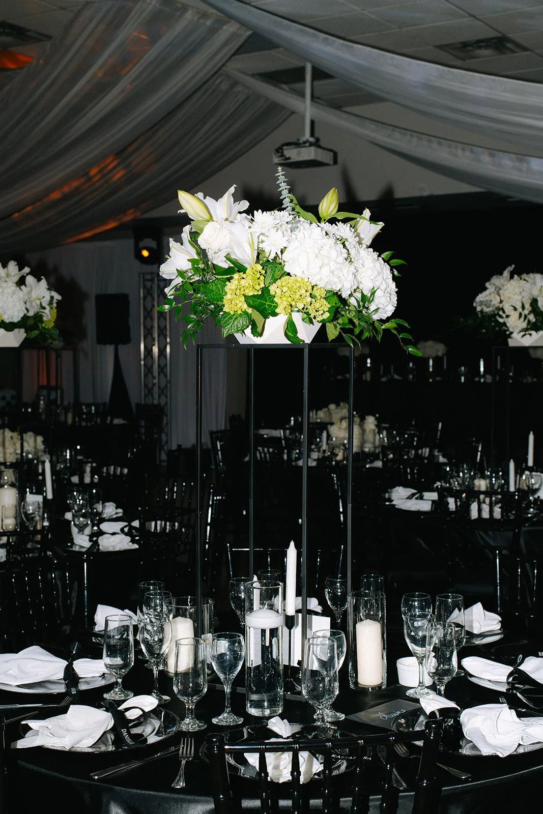 Elegant event table with tall black metal stand holding white floral centerpiece, surrounded by black chairs, white napkins, and glassware, in a dimly lit venue with draped ceiling