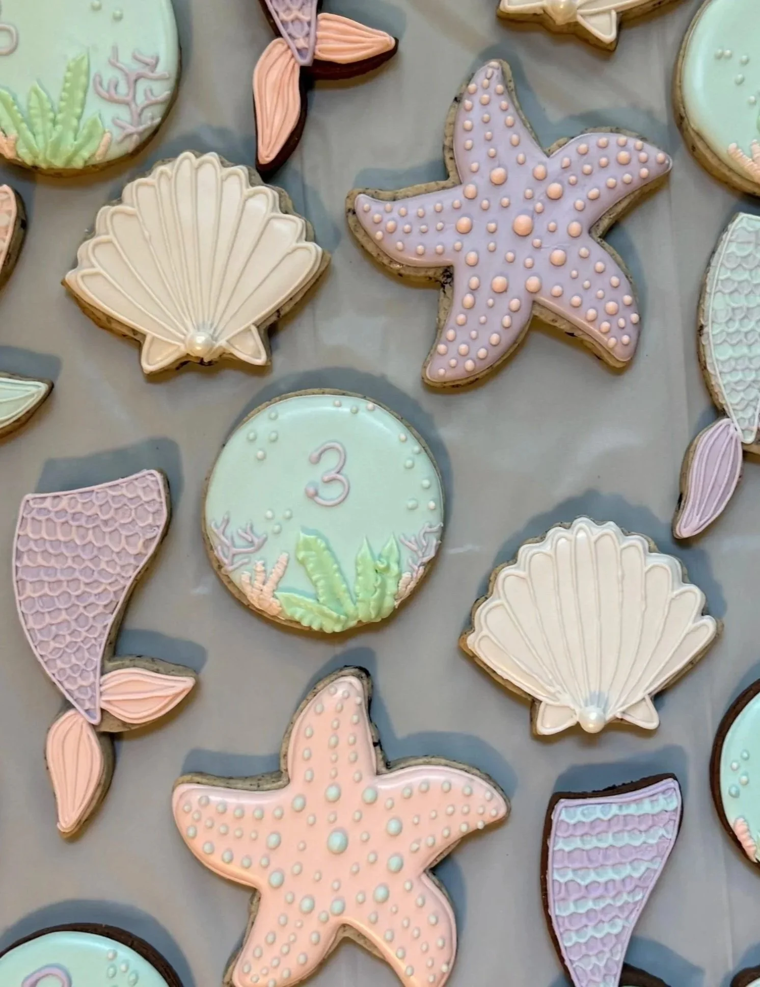 Colorful decorated cookies shaped like seashells, starfish, and mermaid tails with pastel pink, mint green, lavender, and peach icing, some with polka dots and textured patterns, on a gray surface.