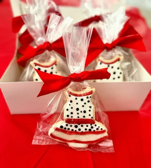 Cookies decorated as black and white polka dot dresses, individually wrapped in clear plastic with red bows, arranged in a white box on a red surface.