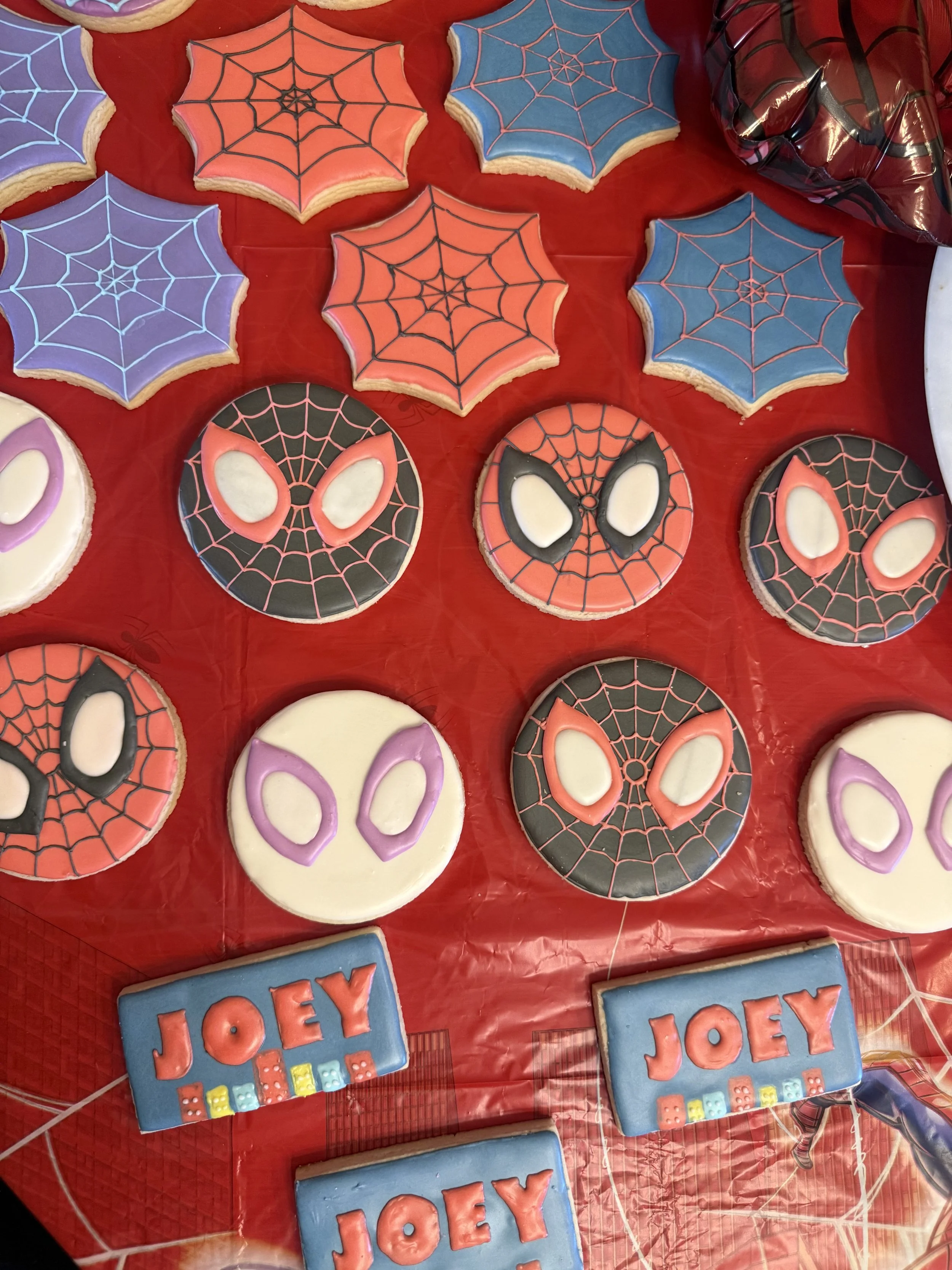 Decorative cookies with spider web and superhero face designs, with some labeled 'JOEY' on a red tablecloth.