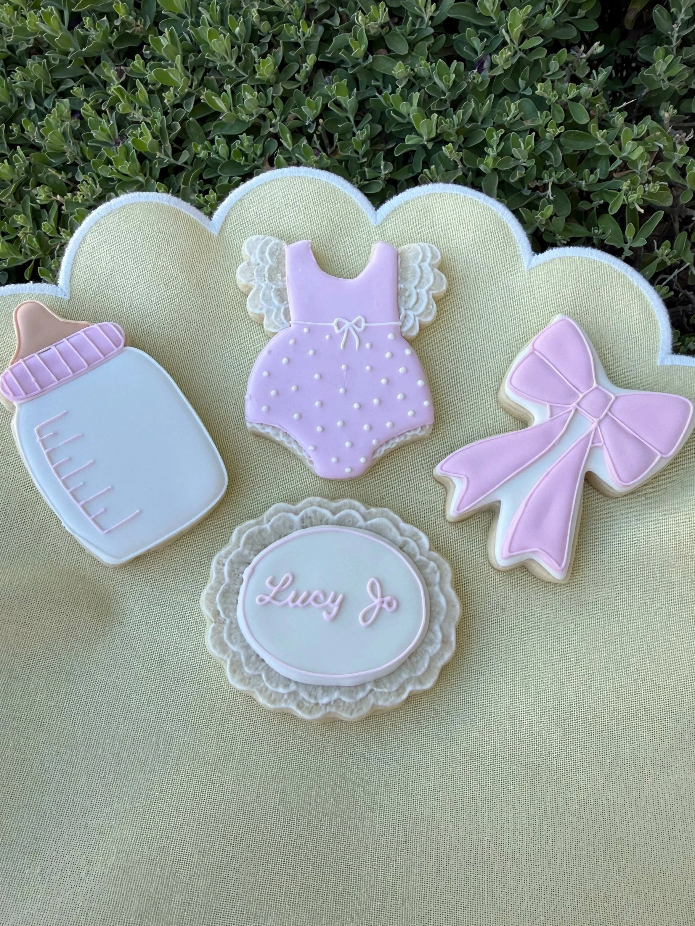 Decorative cookies in pastel pink, purple, and white colors with designs including a baby bottle, a dress with angel wings, a bow, and the name Lucy Jo on a scalloped edge plaque, arranged on a light yellow fabric with green bushes in the background.