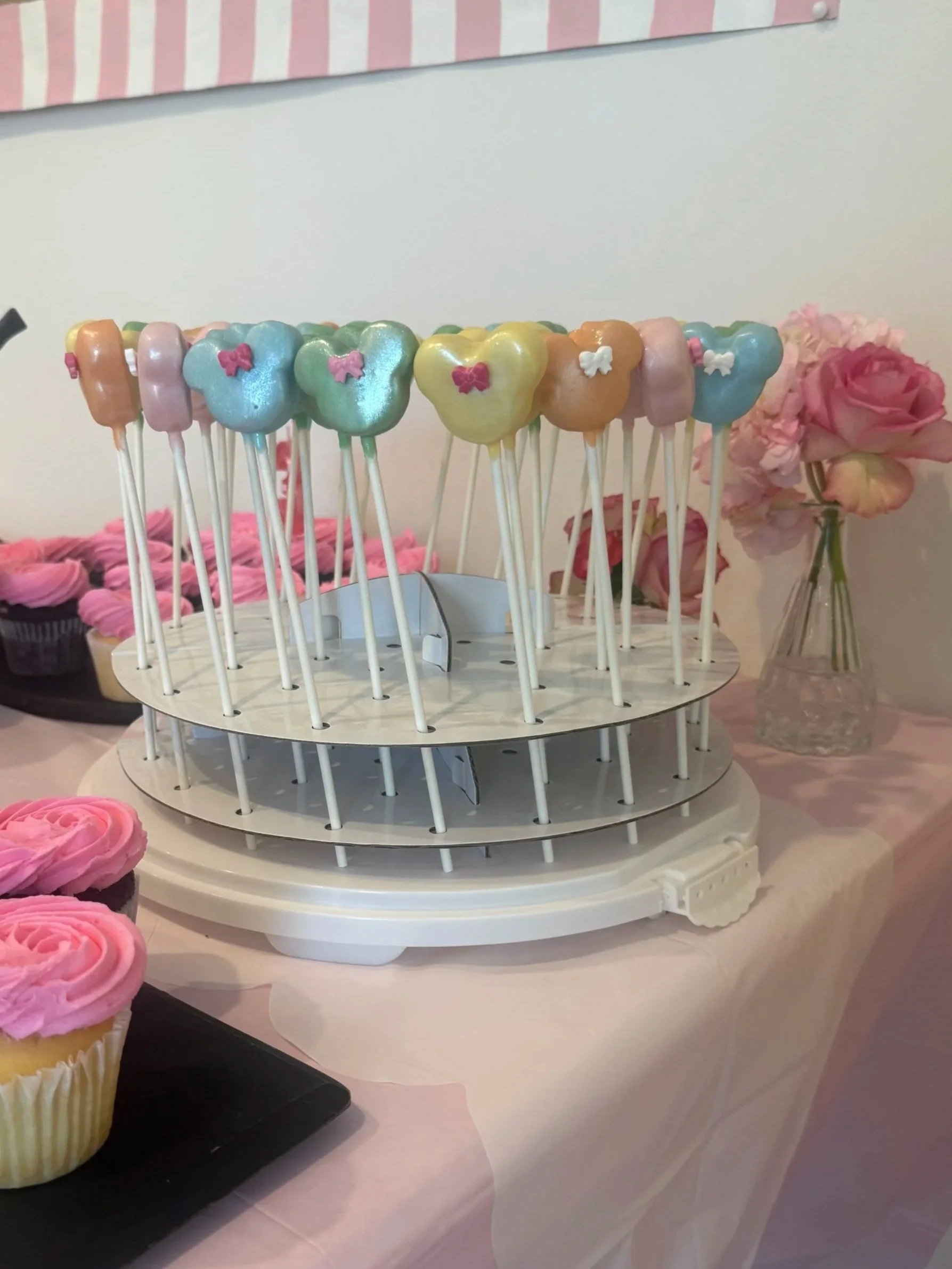 Colorful cake pops on a white display stand, decorated with small bows, in pastel shades of pink, blue, yellow, orange, and green, arranged in a circular pattern. A vase with pink flowers is seen in the background.