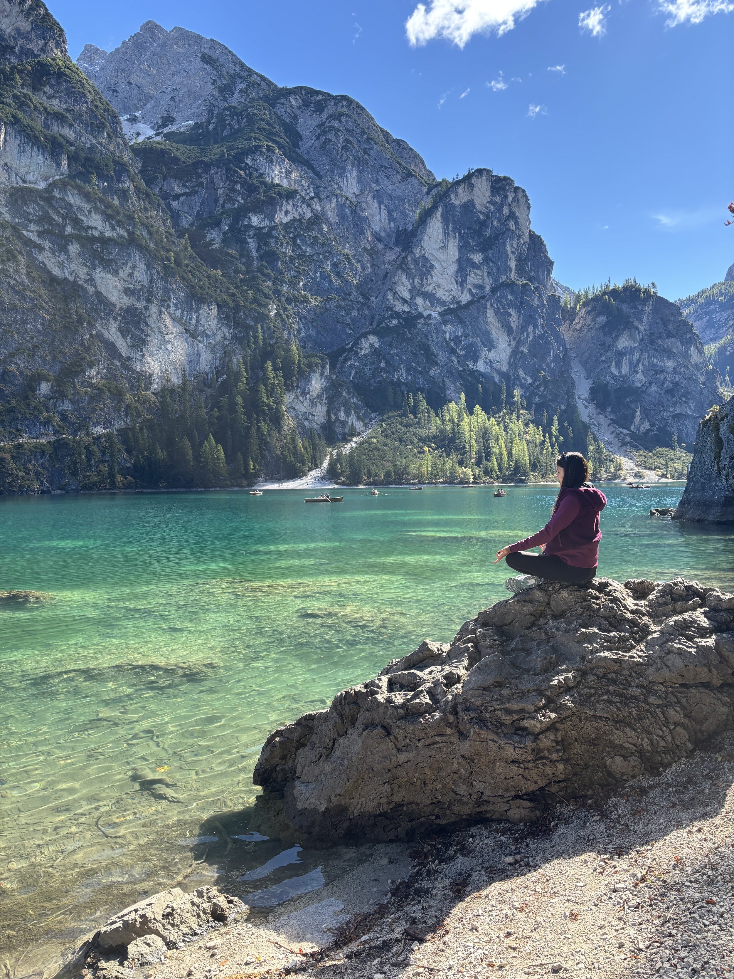 A woman sitting on rocks at the edge of a turquoise lake with mountains and a clear blue sky in the background.