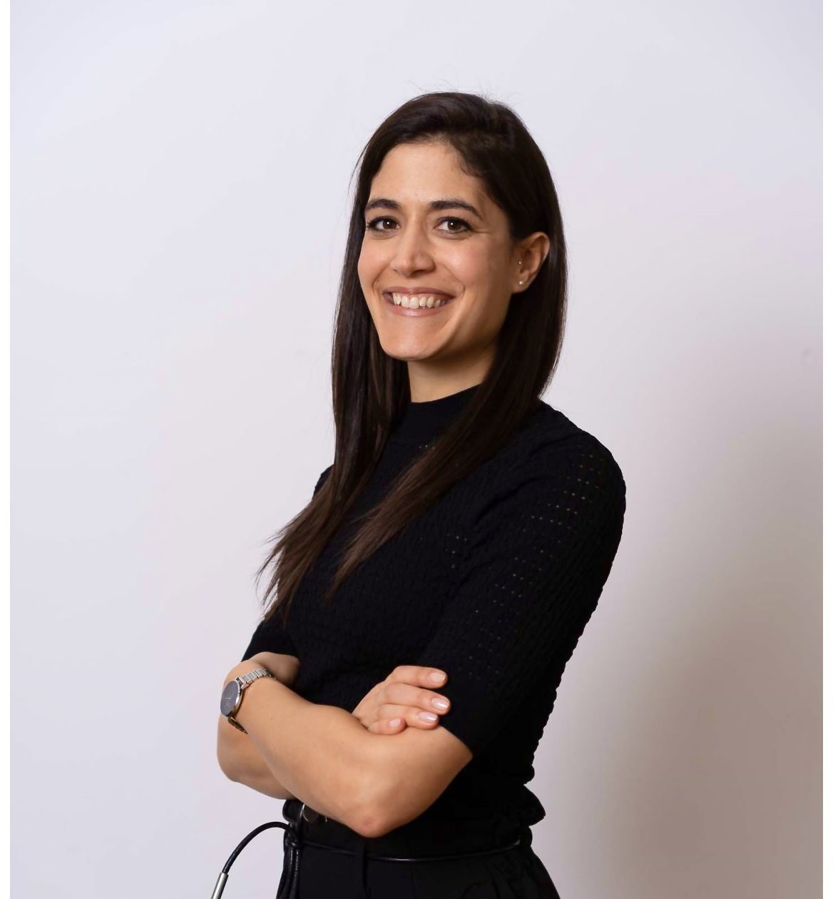 A young woman with long dark hair and a black top, smiling and standing in front of a plain white background with arms crossed.