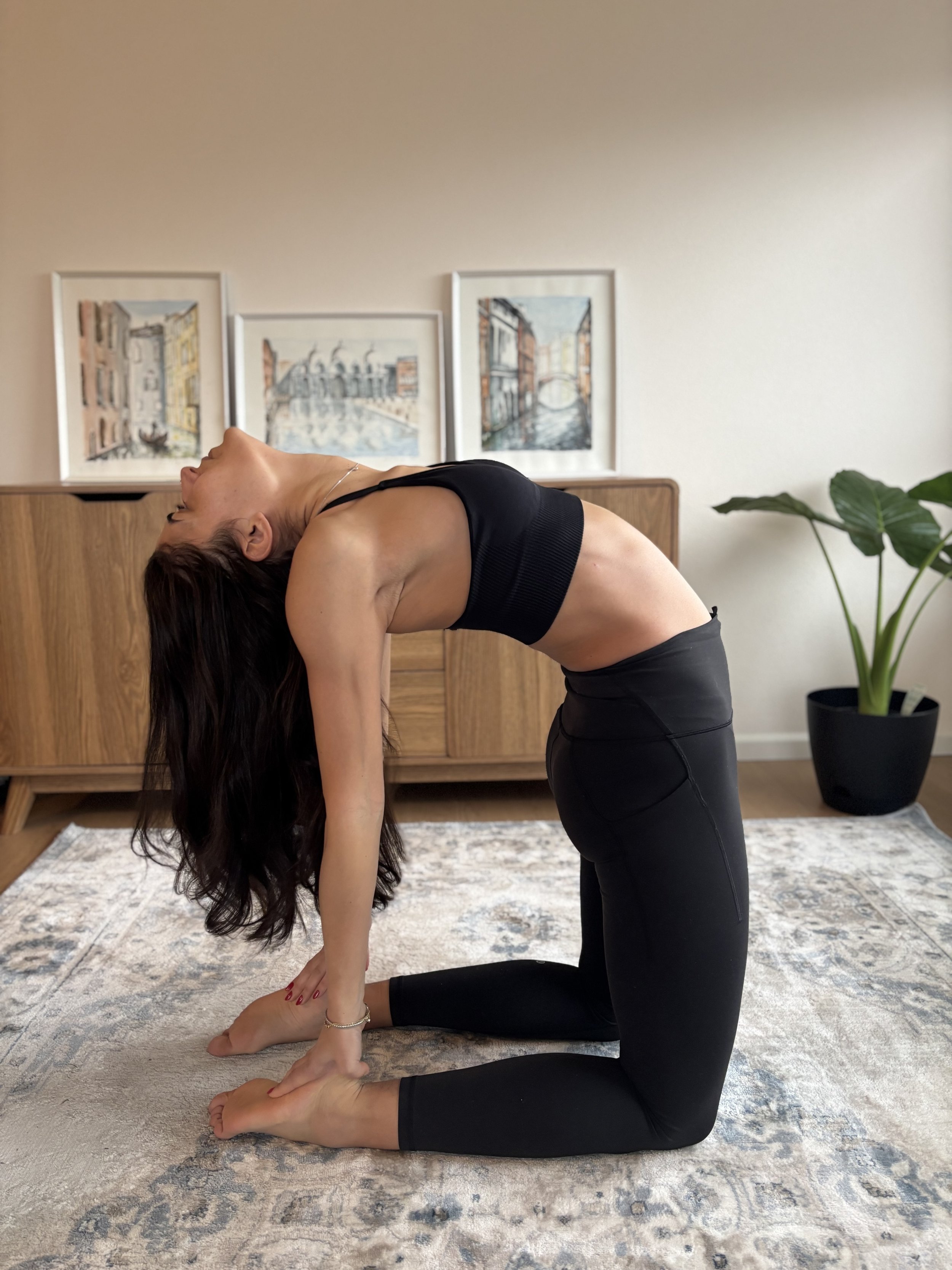 A woman practicing yoga indoors, performing a backbend stretch on a patterned rug, with framed artwork hanging on a wooden cabinet and a large indoor plant nearby.