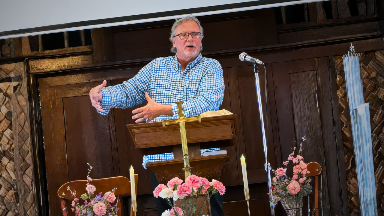 Pastor Tom Crouse wearing a checkered blue and white shirt, standing at a wooden podium with a brass cross, giving a speech or sermon. The setting is a rustic wooden chapel or church with pink and white flowers, candles, and a microphone.