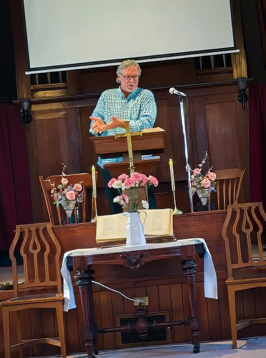 A man speaking at a church pulpit with an open Bible, floral arrangements, and lit candles, with a large projection screen behind him.