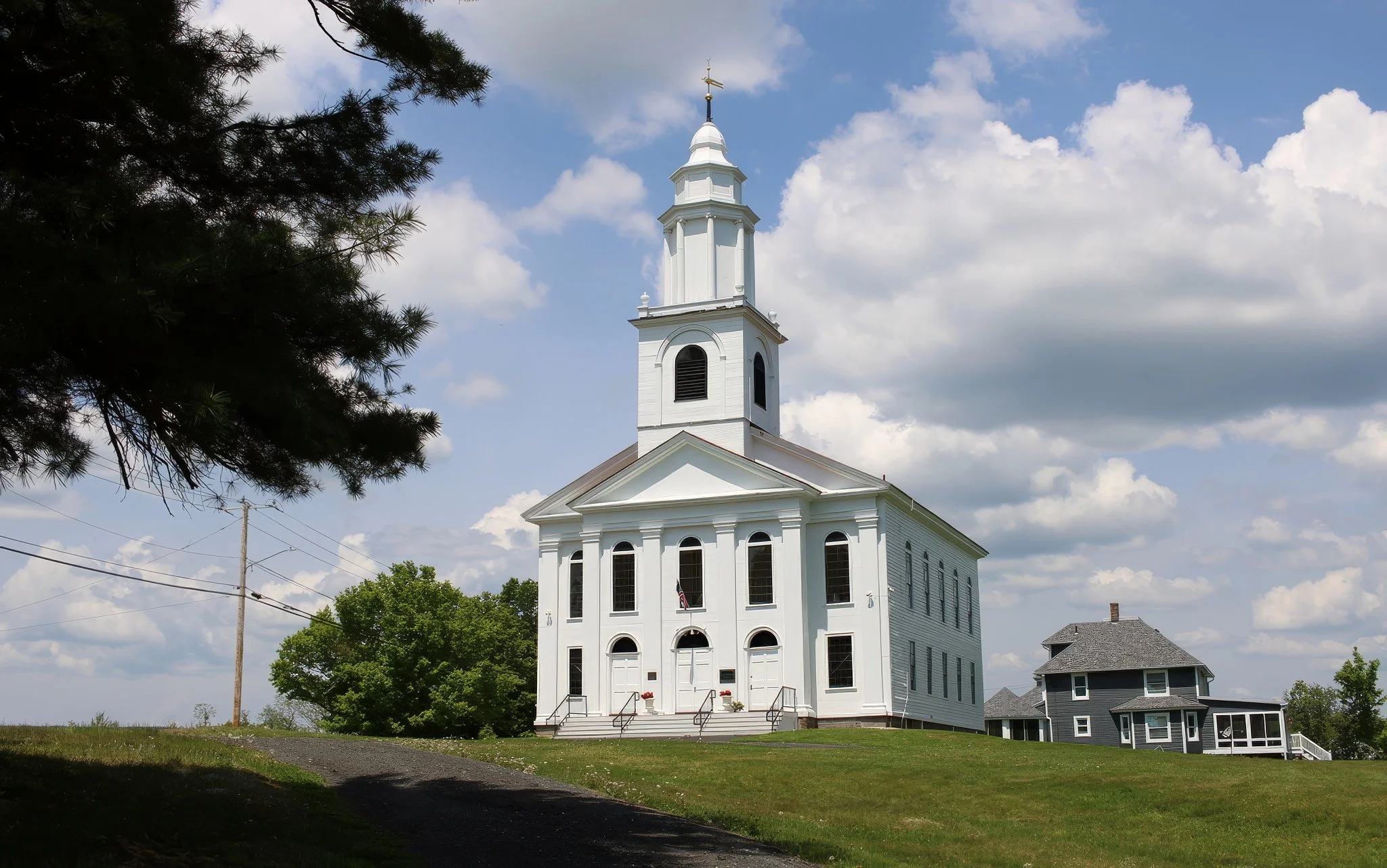 A white church with a steeple and a weather vane on top, set against a partly cloudy sky, with green trees and other houses nearby.