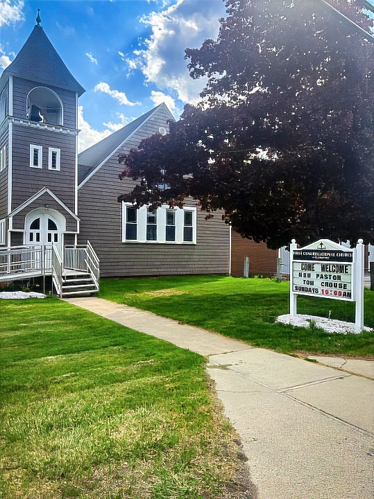 Exterior view of First Congregational Church of Blandford with a gray wooden facade, white trim, a steeple, and a large tree, with a sidewalk leading to the entrance and a sign welcoming the congregation for Sunday services