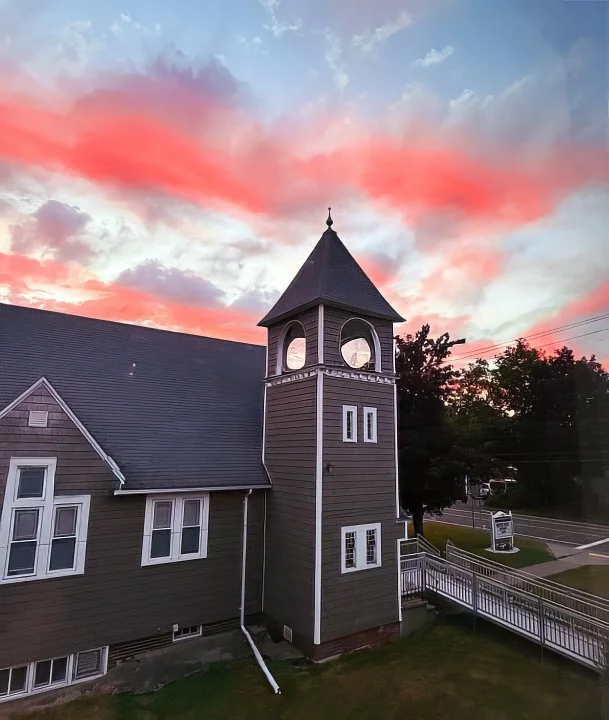 A church building with a tower and clock windows, gray siding, and a pointed roof, set against a colorful pink and blue sunset sky. There are trees, a sidewalk, and electric wires nearby.