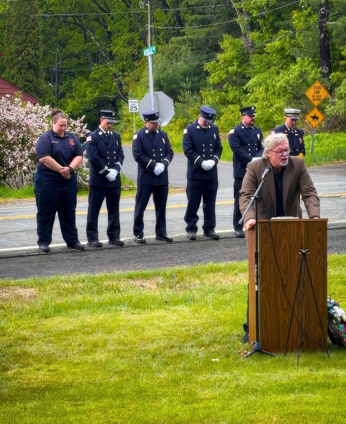 Pastor Tom Crouse is speaking at a podium outdoors, while five people, some in police uniforms, stand behind him with bowed heads in a moment of silence. The scene is on a grassy area near the street with trees and signs in the background.