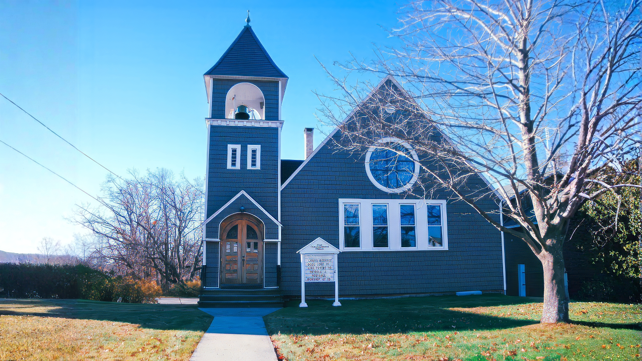 A small church building (First Congregational Church of Blandford) painted dark blue with white trim, a steeple, front steps, and a sign in front, surrounded by a grassy lawn and a leafless tree, under a clear blue sky.