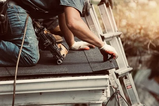 A construction worker in jeans and gloves kneels on a roof, working with roofing materials and tools.
