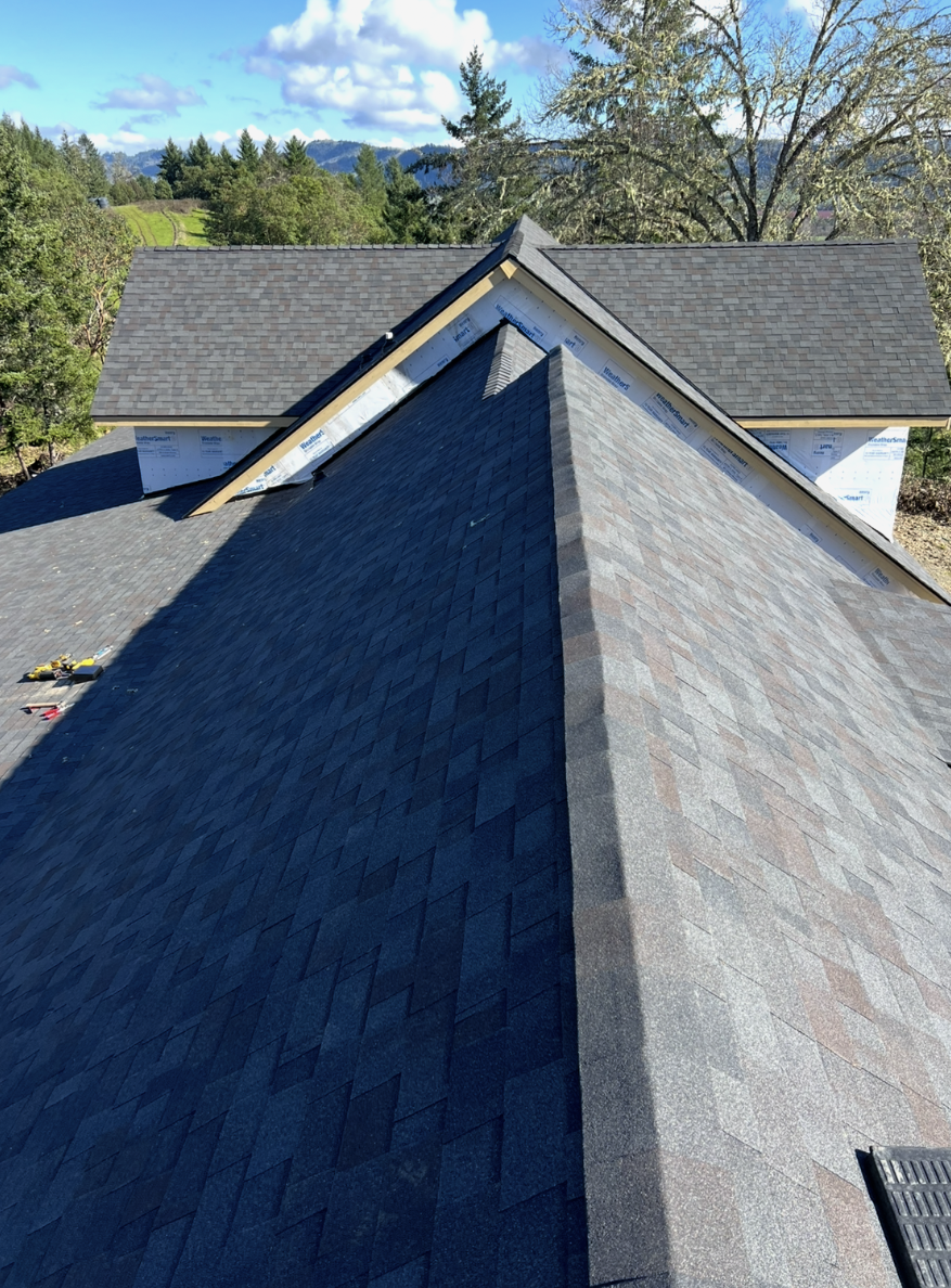 A view of a house roof under construction with new asphalt shingles, with the completed roof visible in the background, set in a scenic area with trees and distant mountains.