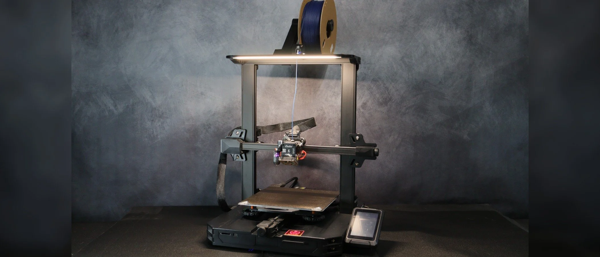 A 3D printer with a spool of blue filament mounted on top, situated against a gray background, with a control touchscreen on the side.