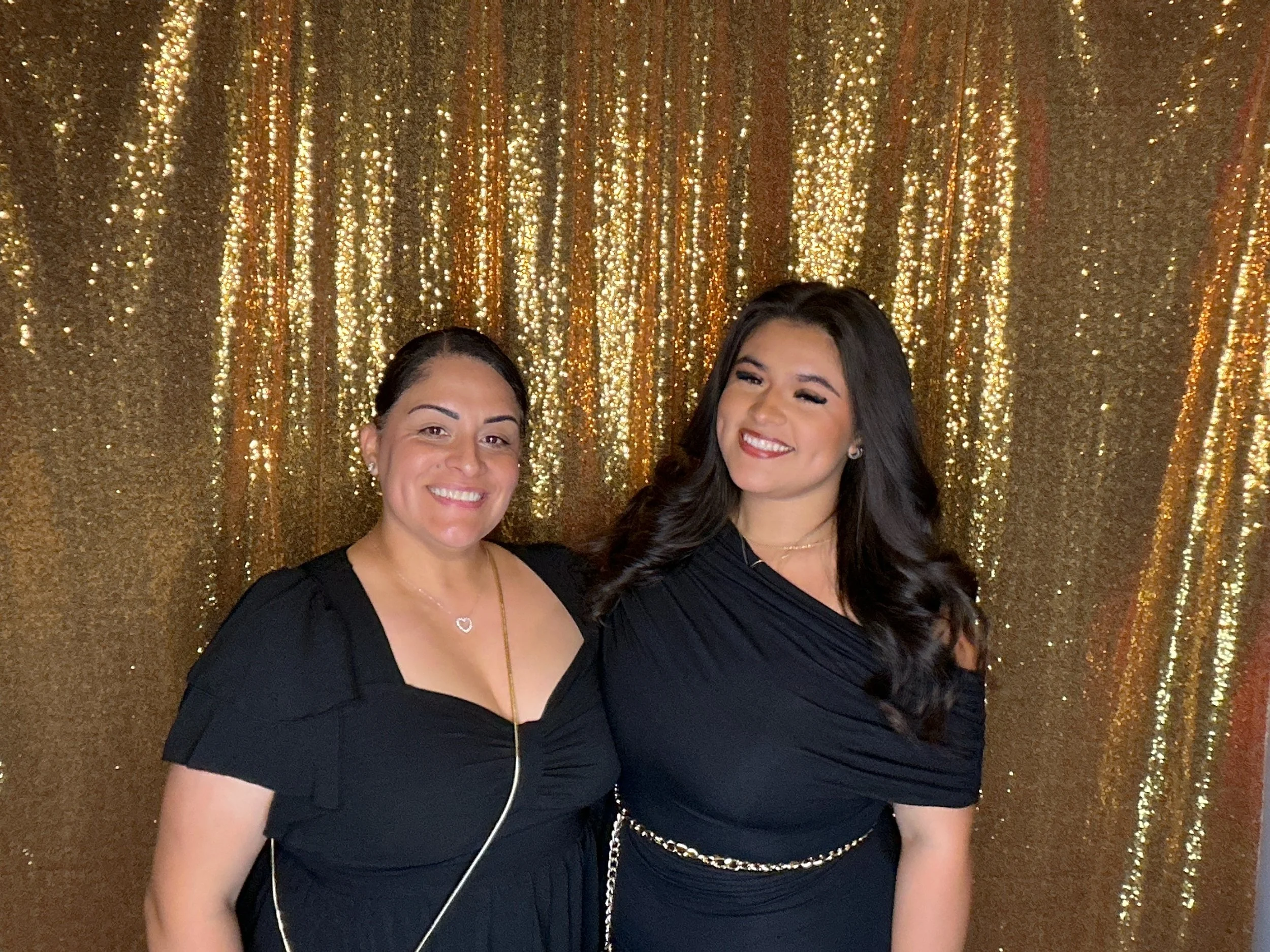 Two women dressed in black, smiling, standing in front of a gold, glittery curtain.