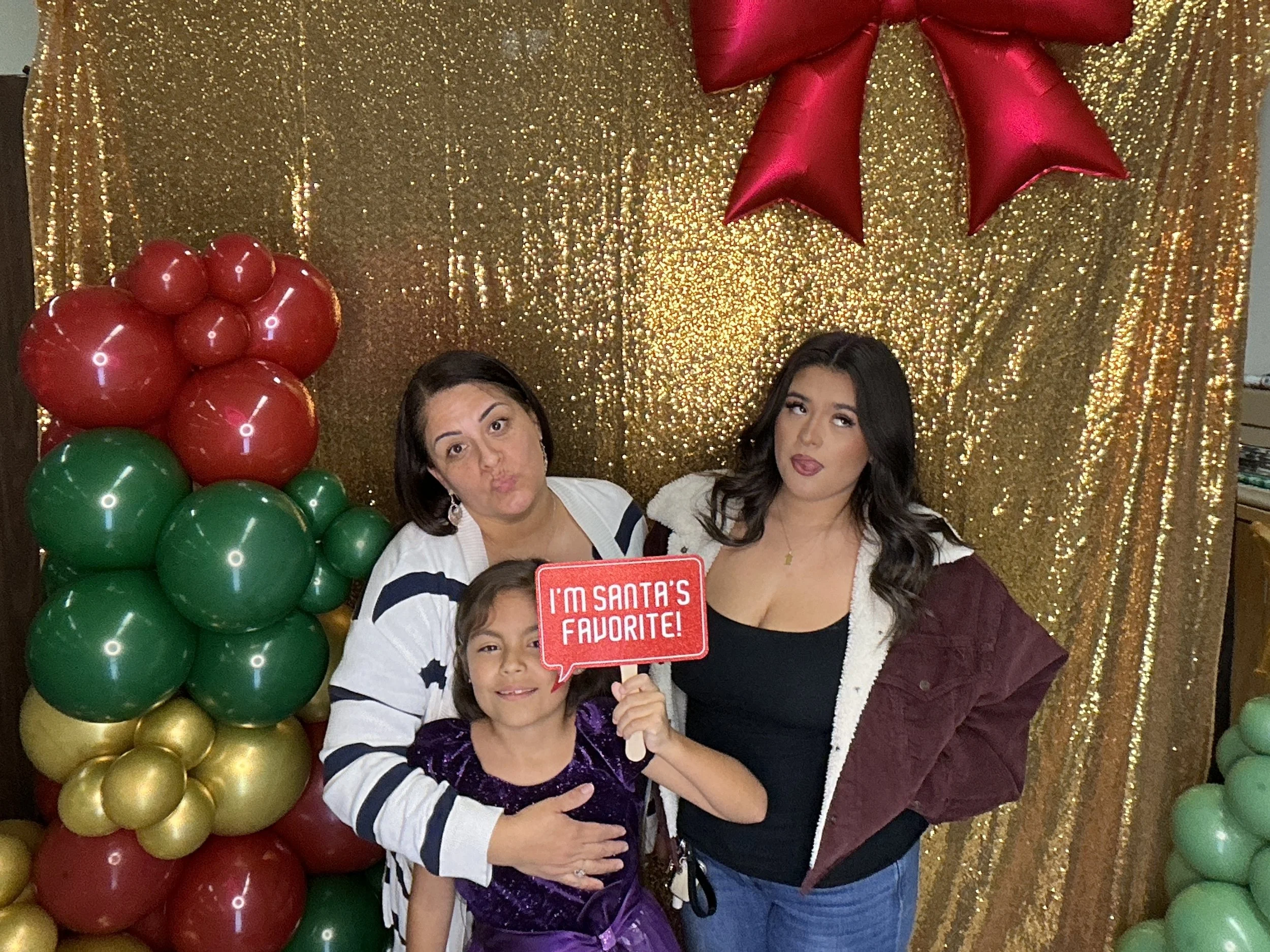 Three women and one girl posing in front of a gold sequin backdrop with red, green, and gold balloons. One woman is holding a red sign that says "I'm Santa's Favorite!" The women and girl are smiling or making playful faces.
