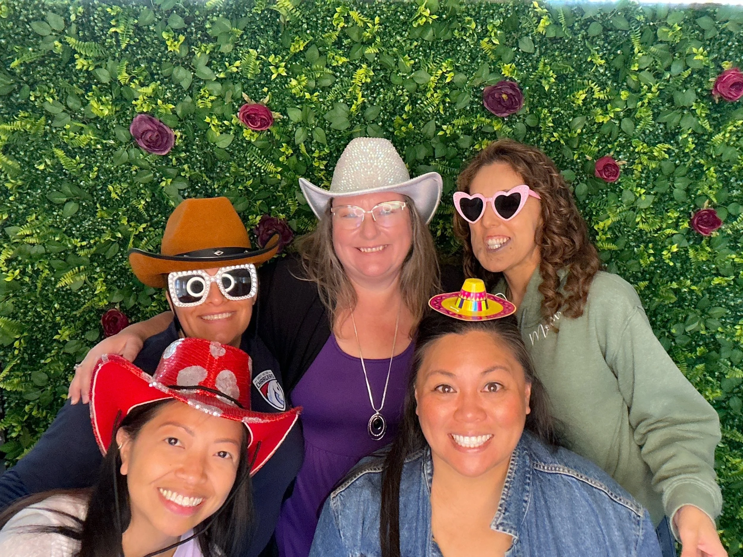 Group of five women posing for a photo in front of a green leafy backdrop with purple roses, wearing fun sunglasses and accessories.