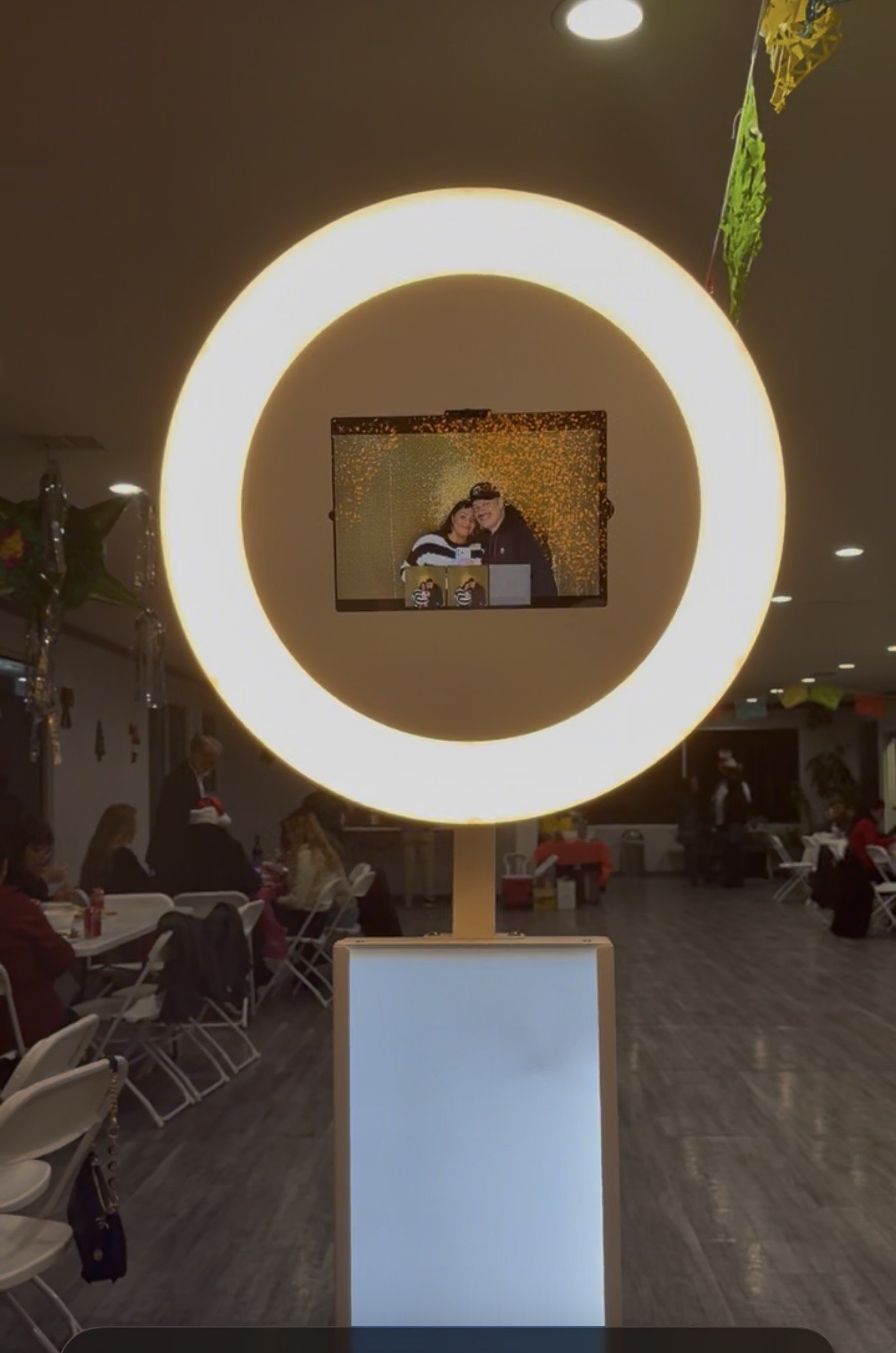Large illuminated circular display with a small screen showing a couple, in a gathering space with people seated at tables.