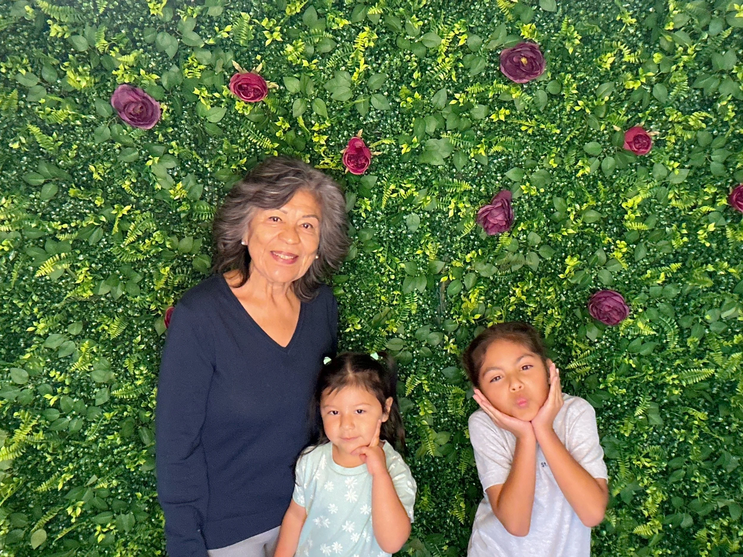 Four women, one older and three young girls, posing together in front of a lush green leafy wall with purple flowers. The older woman has gray hair and is wearing a dark blue top. The three girls have different hairstyles and are making playful faces, with one girl holding her face with her hands and the others making gestures.