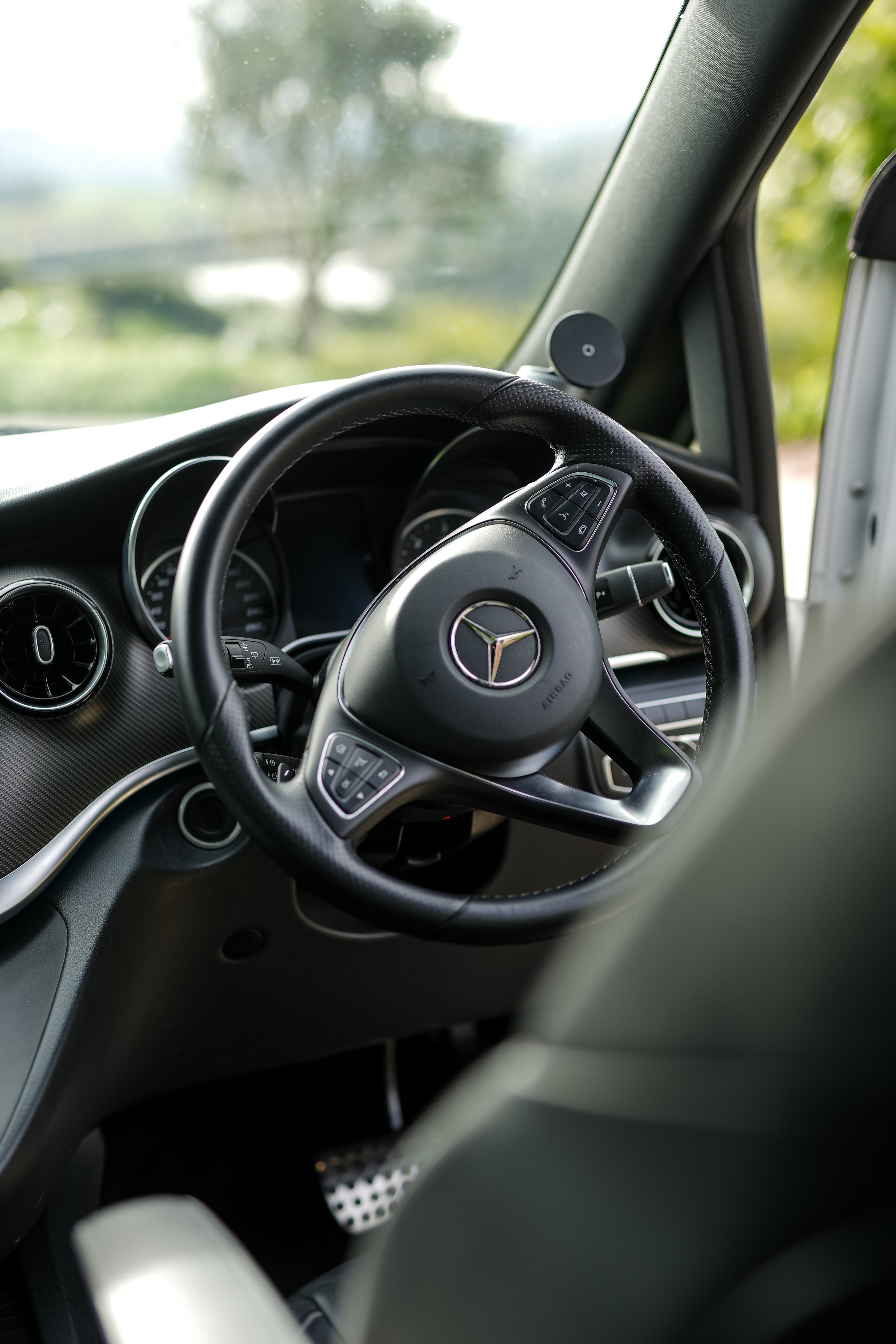 The interior of a Mercedes-Benz vehicle, showing the steering wheel with the Mercedes-Benz logo, dashboard, and part of the windshield with a blurred outdoor background.