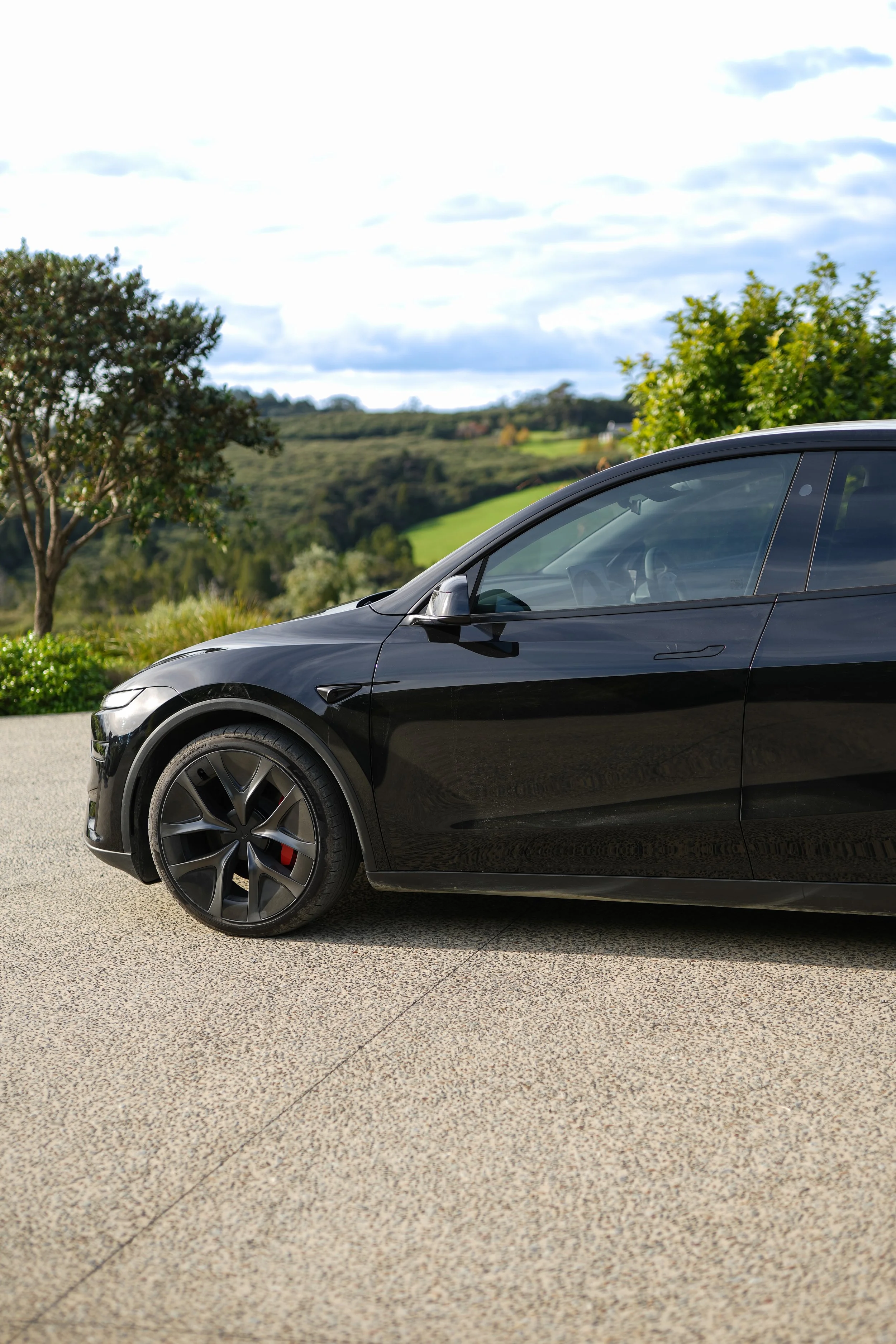 A close-up of a black Tesla Model 3 electric car parked on a light-colored textured driveway with trees and rolling hills in the background and partly cloudy blue sky.