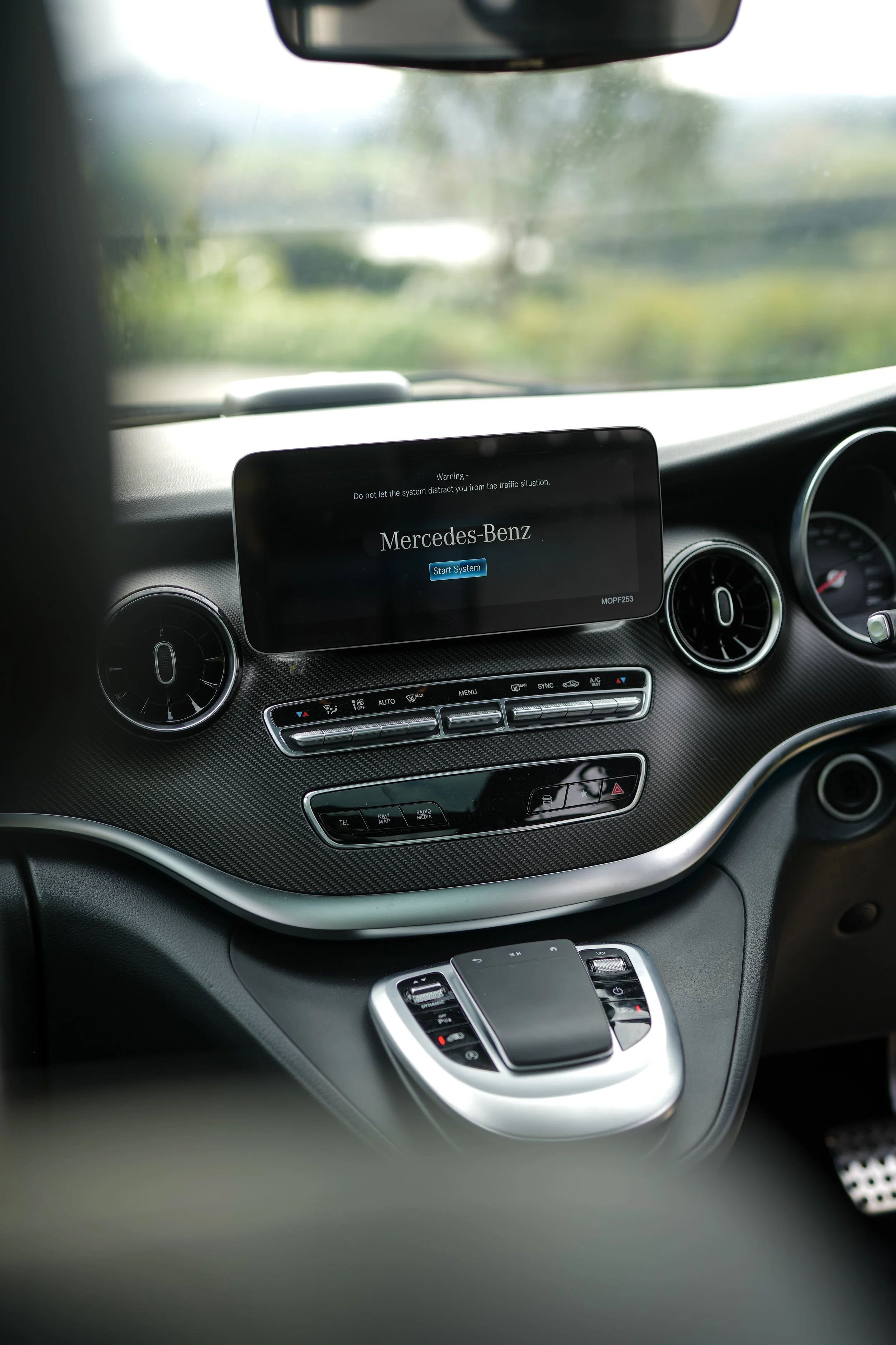 The interior of a Mercedes-Benz vehicle, with focus on the dashboard and central control panel. The dashboard displays a screen with the Mercedes-Benz logo and a start system button. Air vents and climate control buttons are visible below the screen. The image is taken from the passenger side, looking towards the front windshield showing a blurry outdoor scene.
