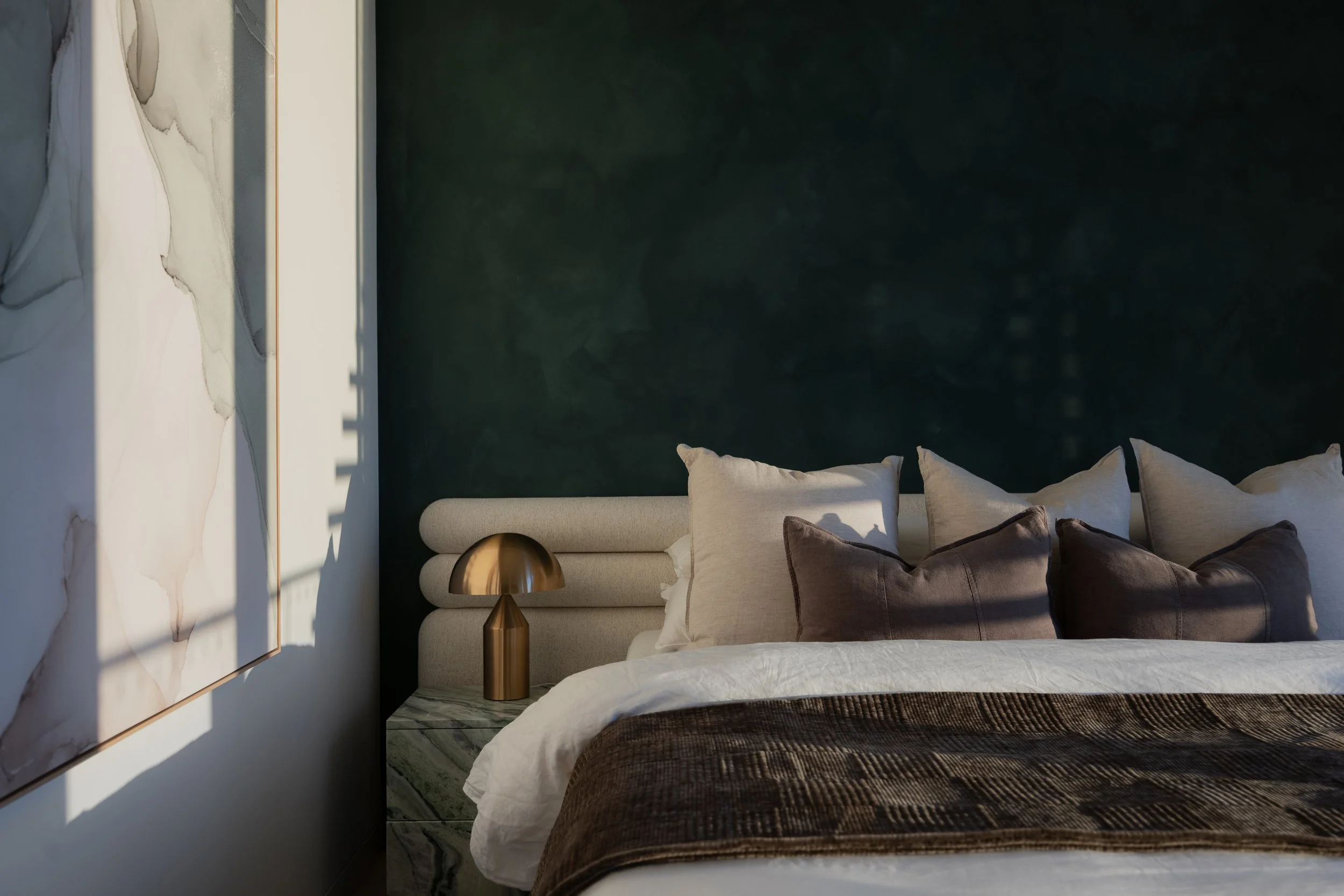 A modern bedroom with a large bed covered in white and brown bedding, against a deep teal wall. There are multiple pillows on the bed. To the left of the bed is a bedside table made of marble with a gold-colored lamp that has a domed shade. On the wall above the bed is a large abstract art piece with neutral tones and flowing lines.