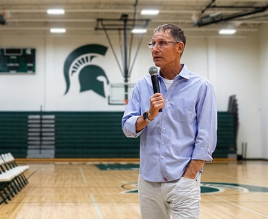 Man speaking into a microphone in a gymnasium with a Spartan logo on the wall.