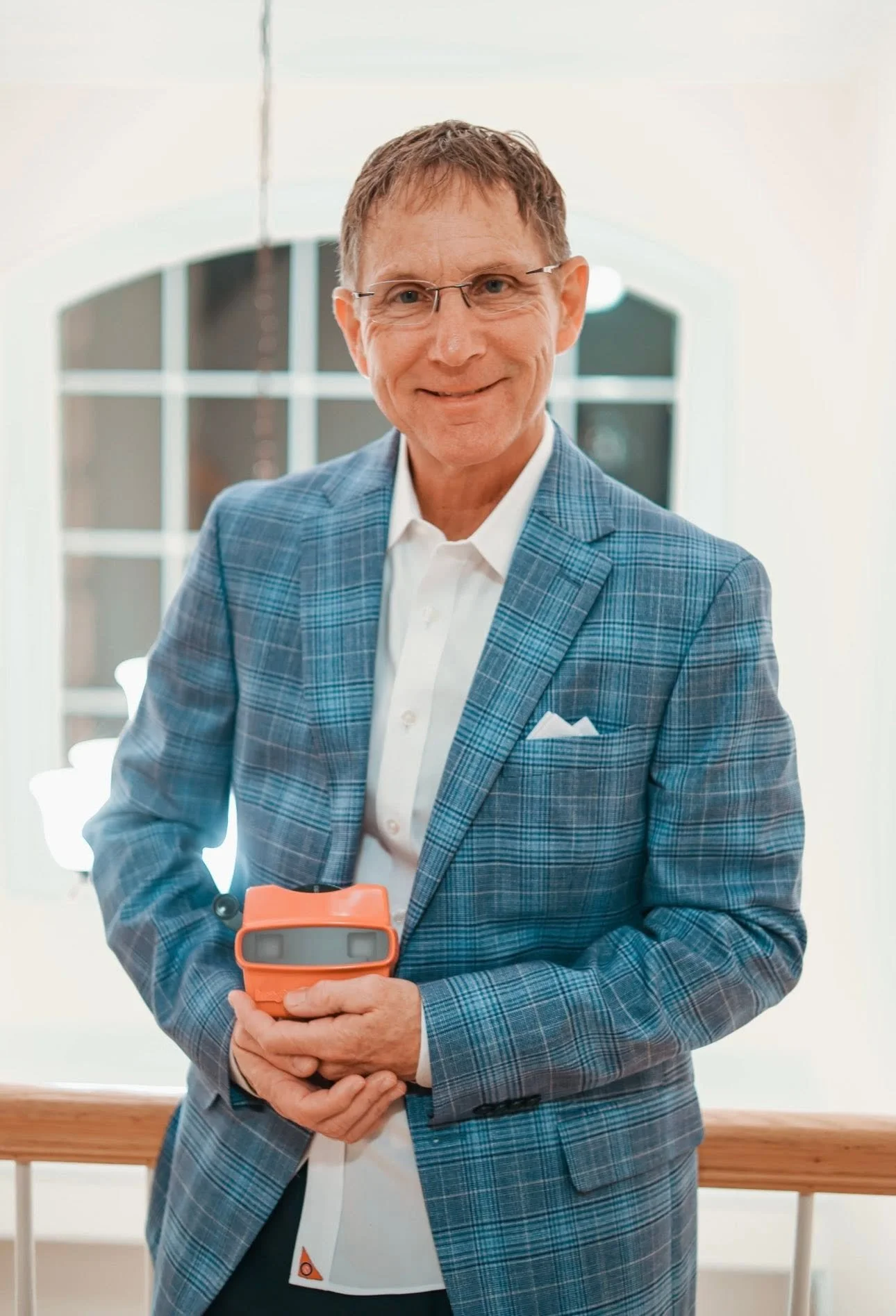 Michael J. Bowen wearing glasses, and sporting a blue plaid blazer and white shirt, is holding a red viewfinder device. He is smiling and standing in a well-lit indoor space with large window panes in the background.