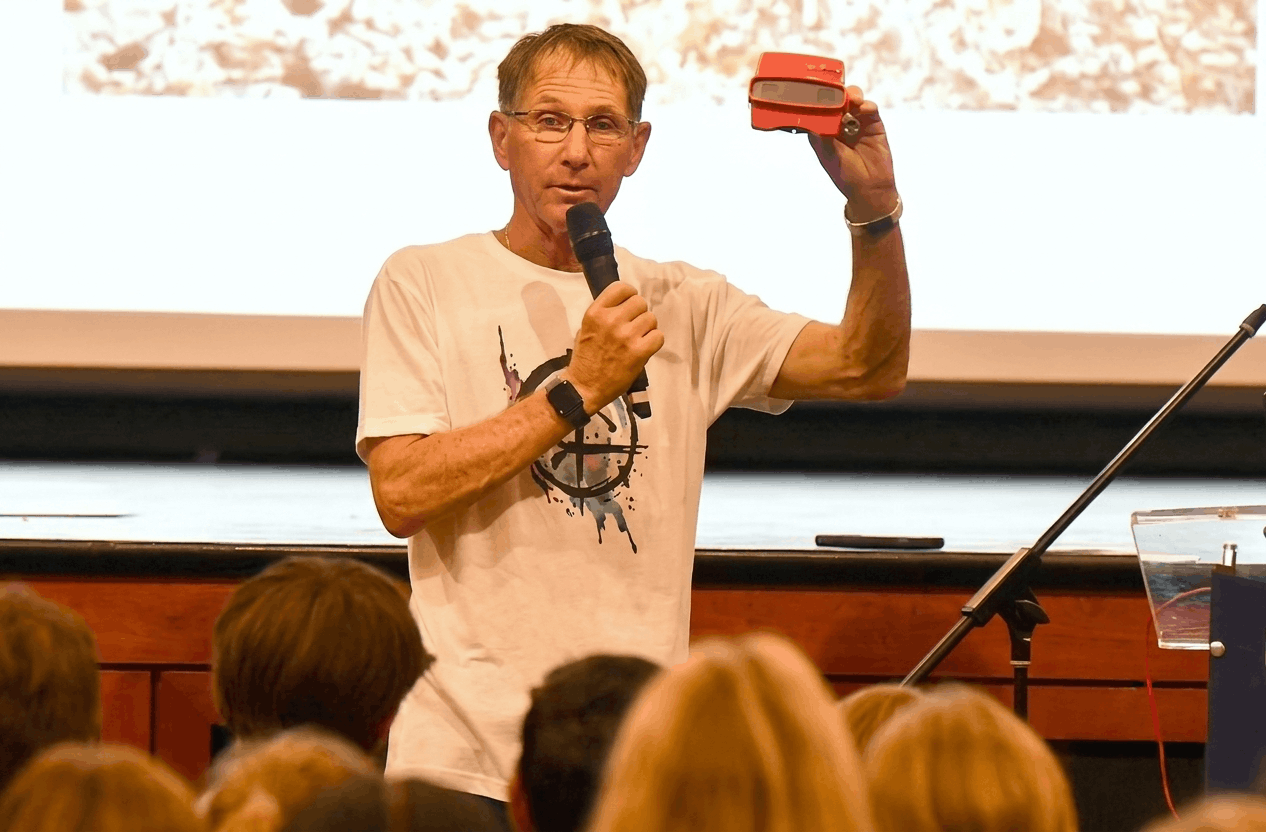 Michael J. Bowen holding a small red viewfinder, speaking to an audience in a lecture hall with a projection screen in the background.