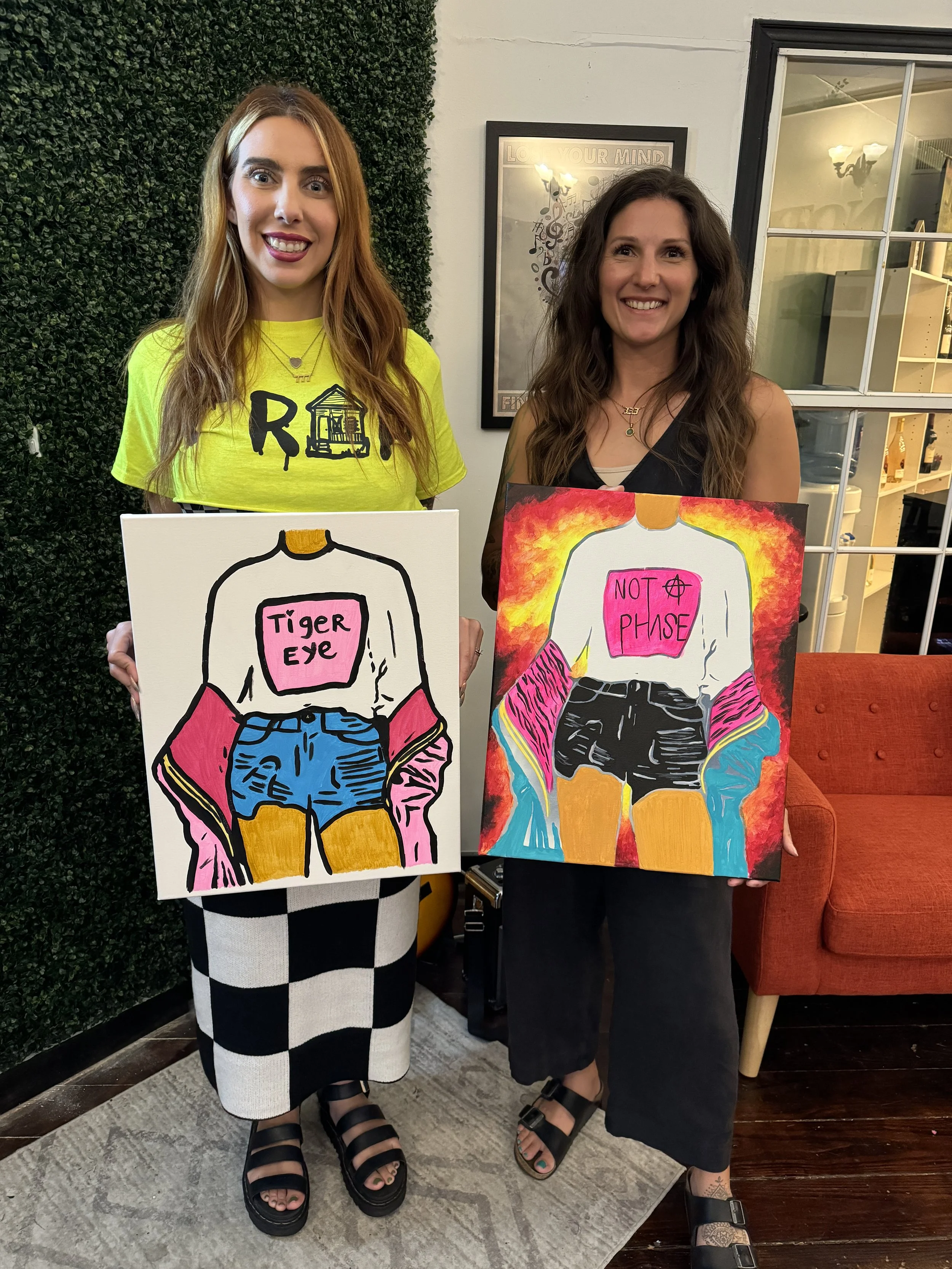two women standing smiling holding sip and paint party canvases