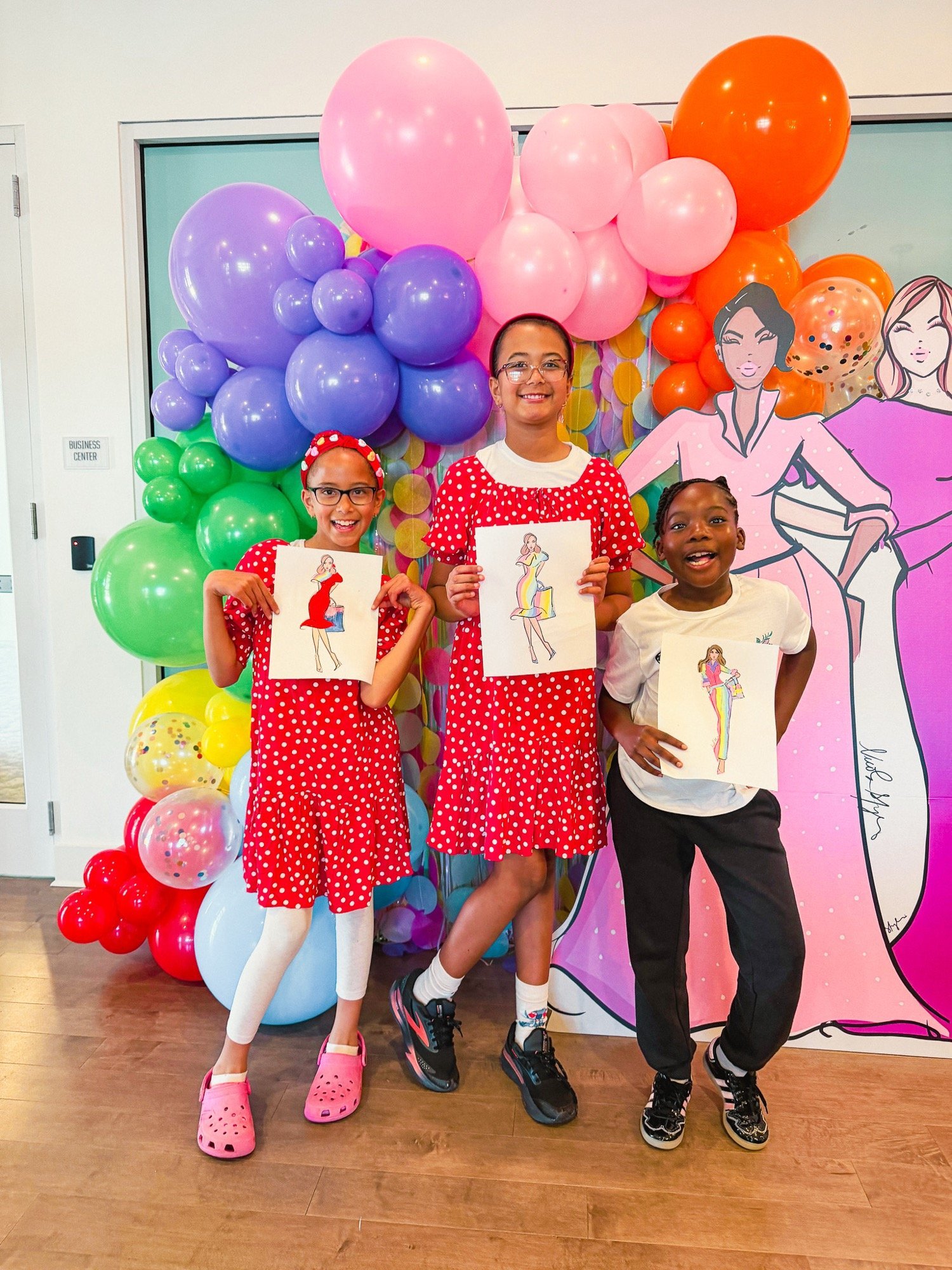 Three children posing in front of a colorful balloon backdrop, holding drawings of women fashionably dressed, with large cartoon-style illustrations of women in the background.