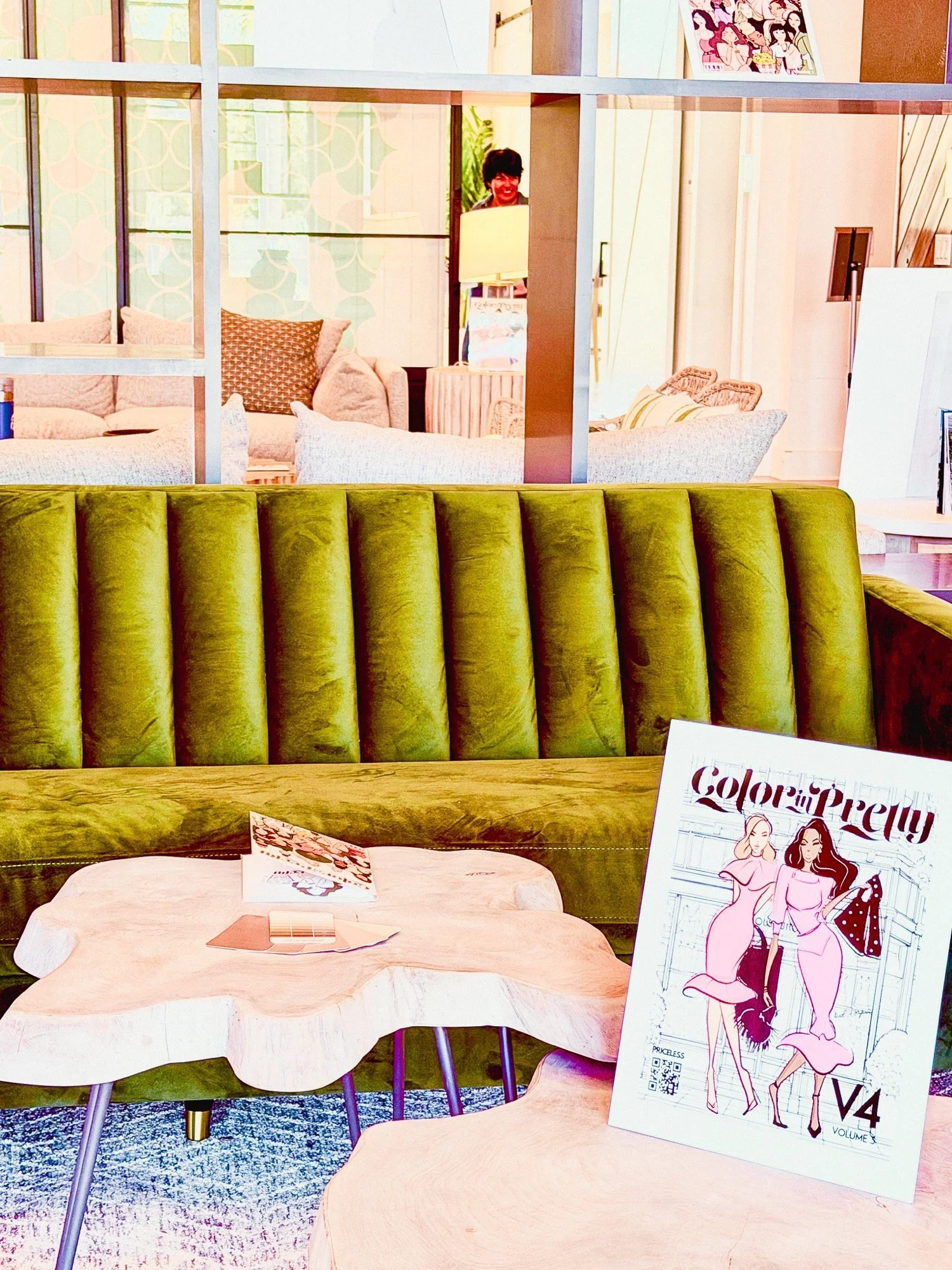 A display of furniture including a green velvet tufted sofa and a round coffee table covered with a blush-colored fabric. On the coffee table, there are promotional materials for 'Color of Pretty' featuring fashion illustrations of women. In the back